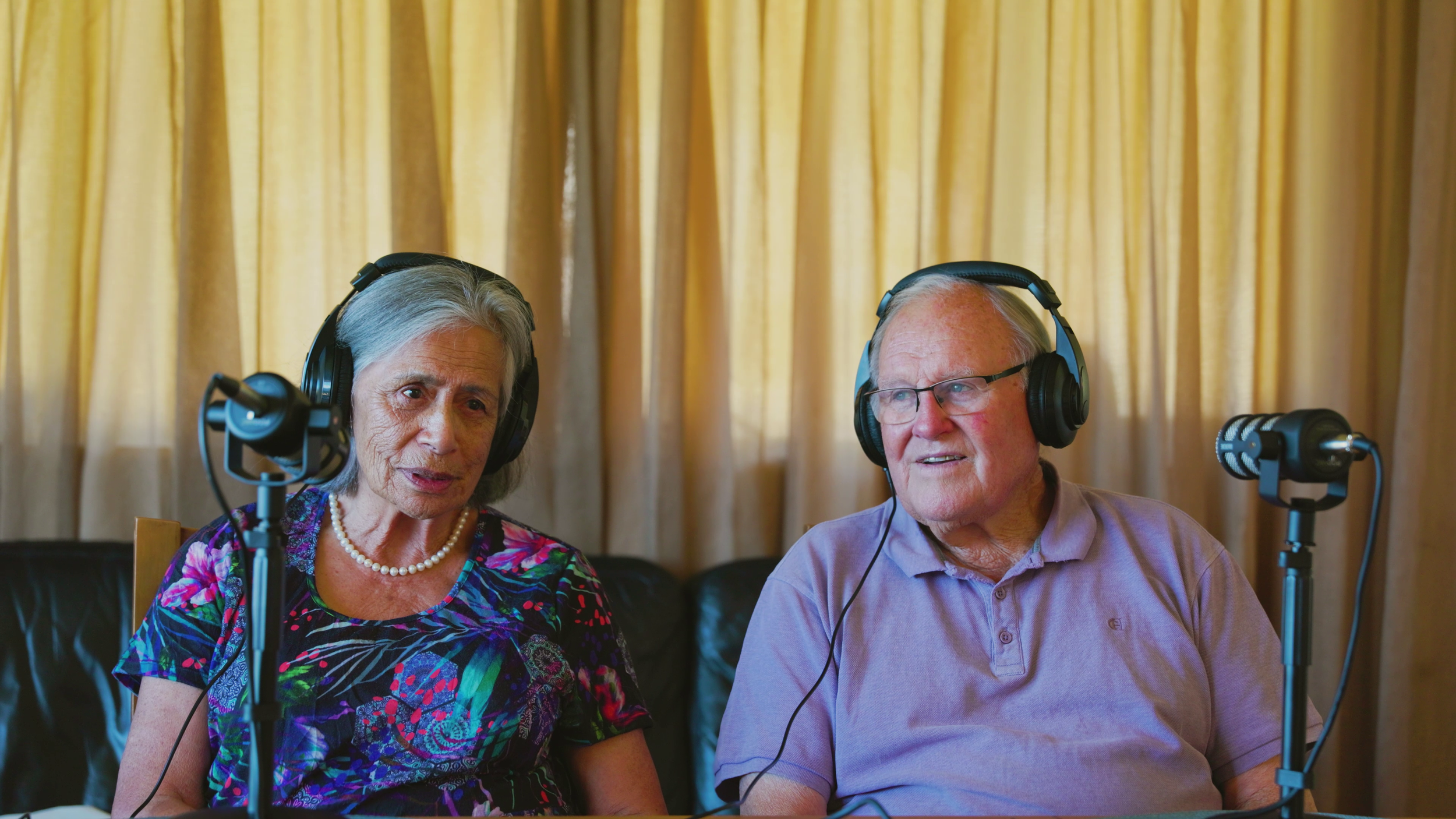 An elderly woman and man sitting at a table wearing headphones, speaking into microphones, with a yellow curtain in the background.