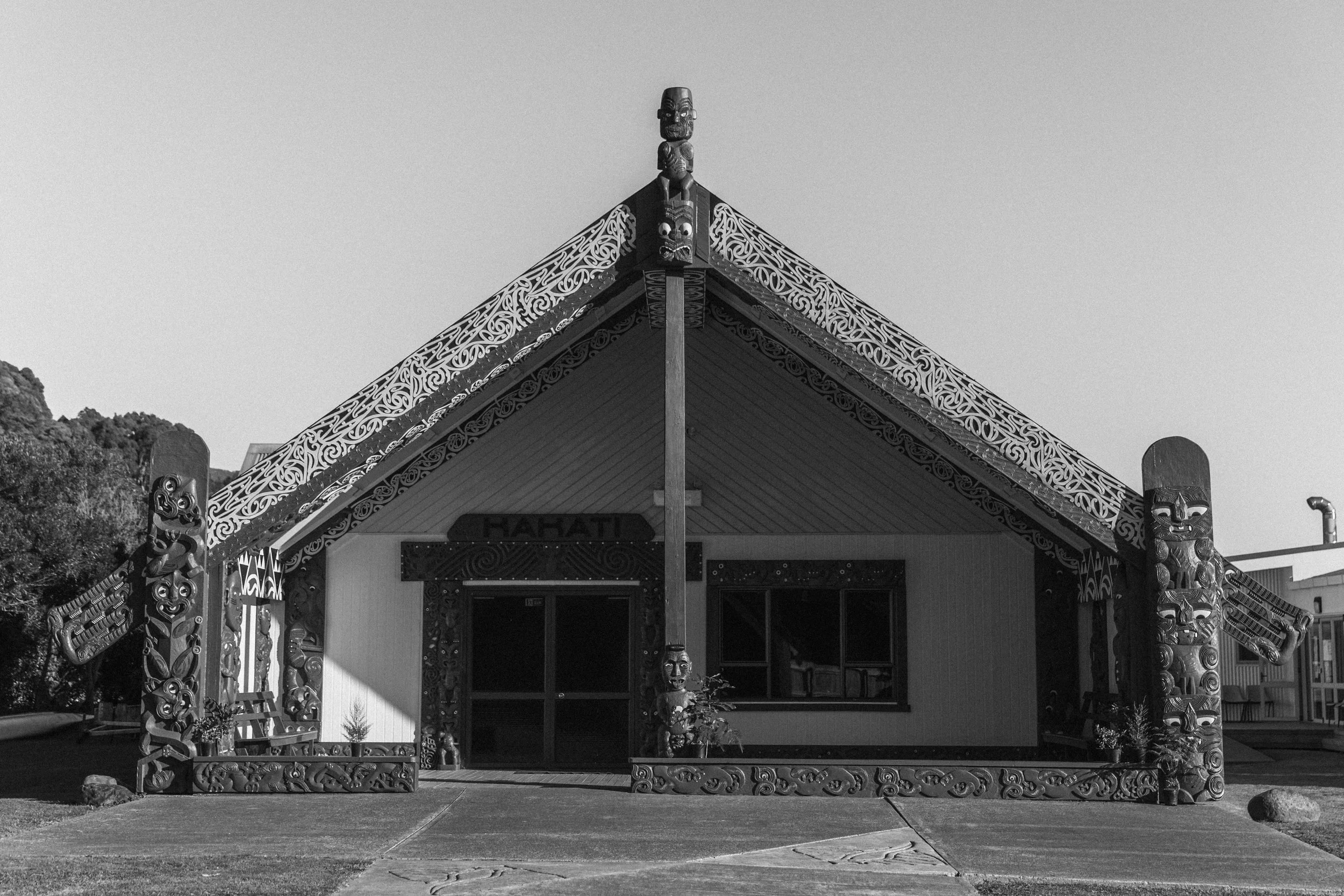 Black and white photo of a traditional Maori meeting house with intricate carvings on the posts and eaves, set against a clear sky.