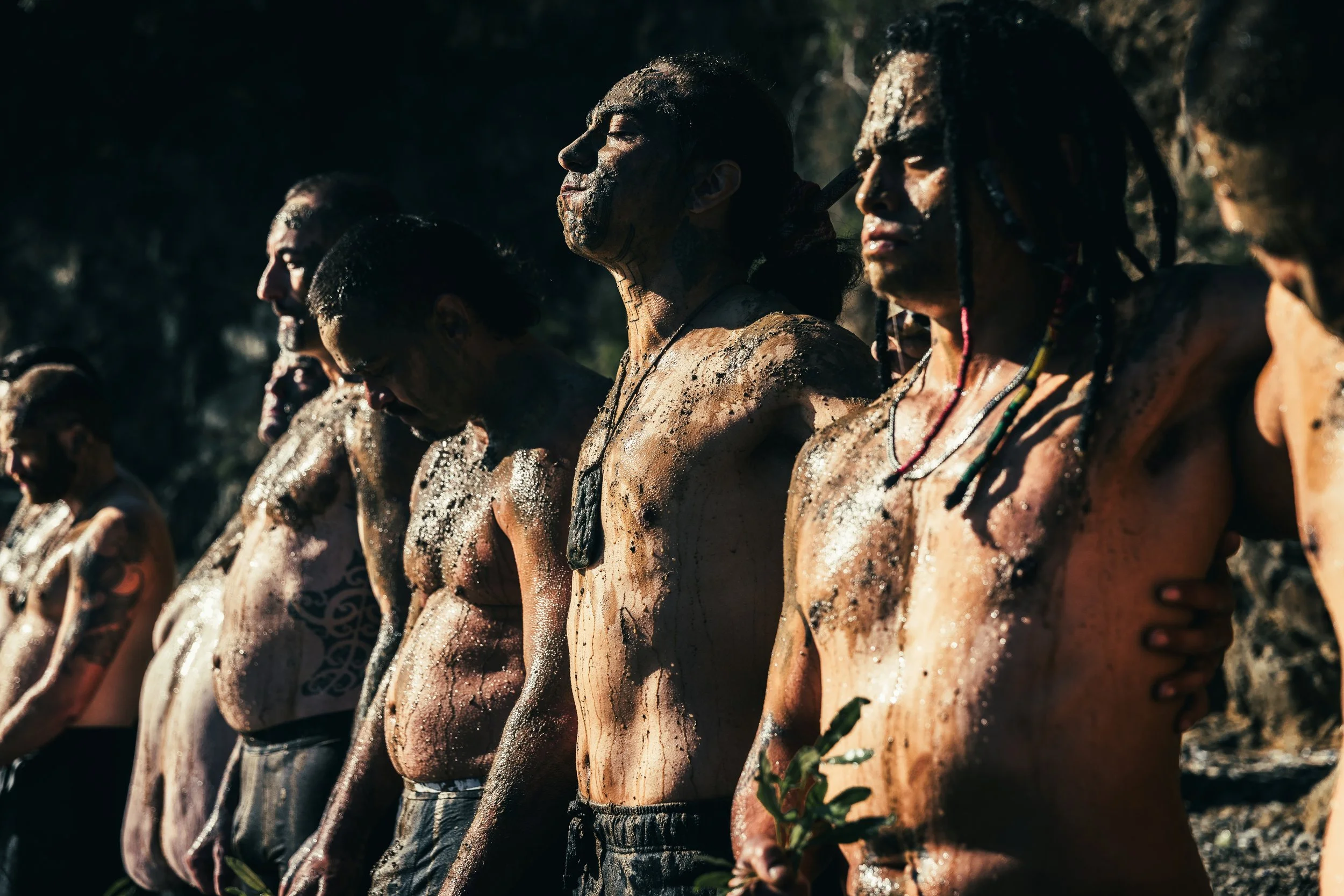A group of indigenous people with traditional body paint and jewelry, standing side by side outdoors during daylight.