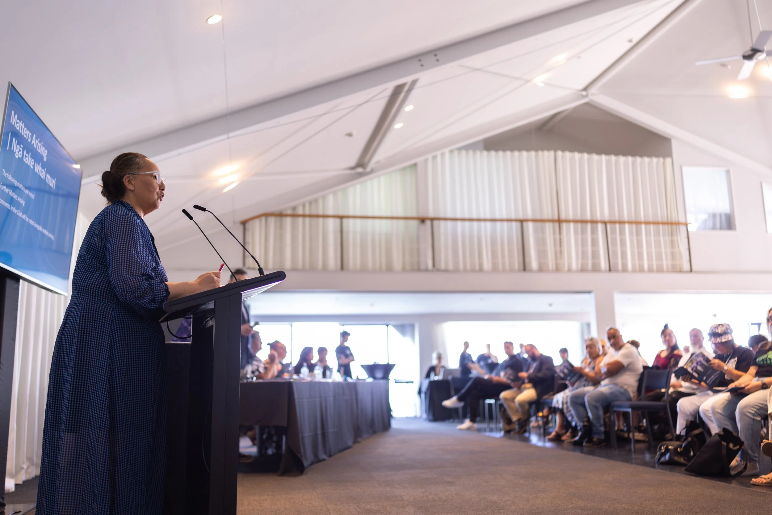 A woman with glasses and her hair in a bun speaking at a podium with microphones during a conference or meeting in a room with high ceilings and an audience seated and listening.