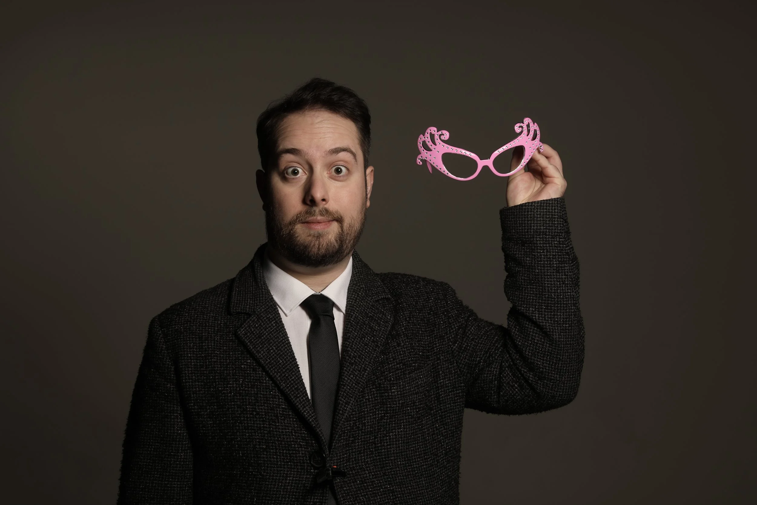 A man with a beard and dark hair wearing a black suit, white shirt, and black tie holding pink decorative glasses, looking at the camera with a surprised expression.