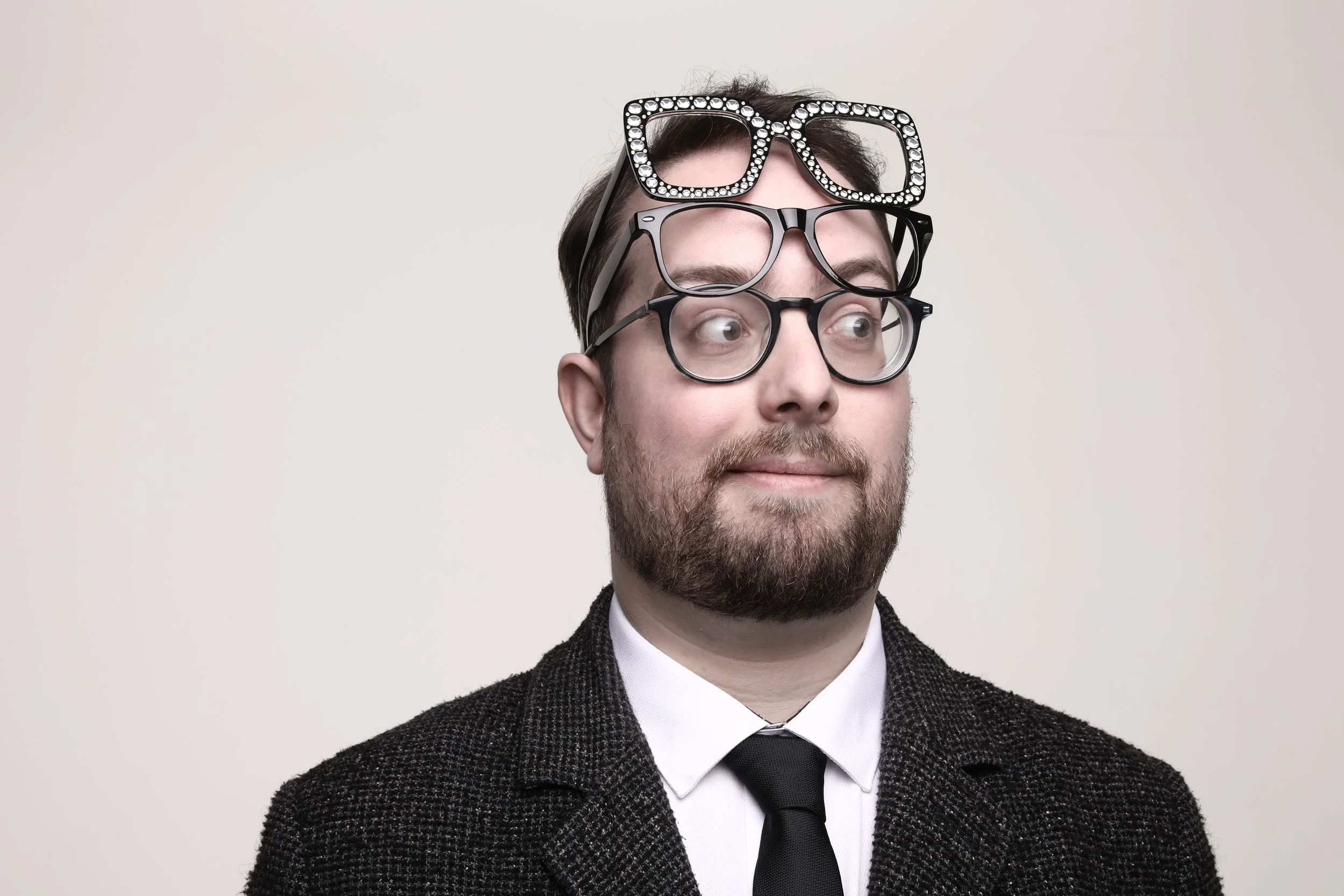 Jake Donaldson Comedian with a beard and dressed in a suit, wearing a shirt and tie, with multiple pairs of glasses stacked on his head, some adorned with rhinestones.
