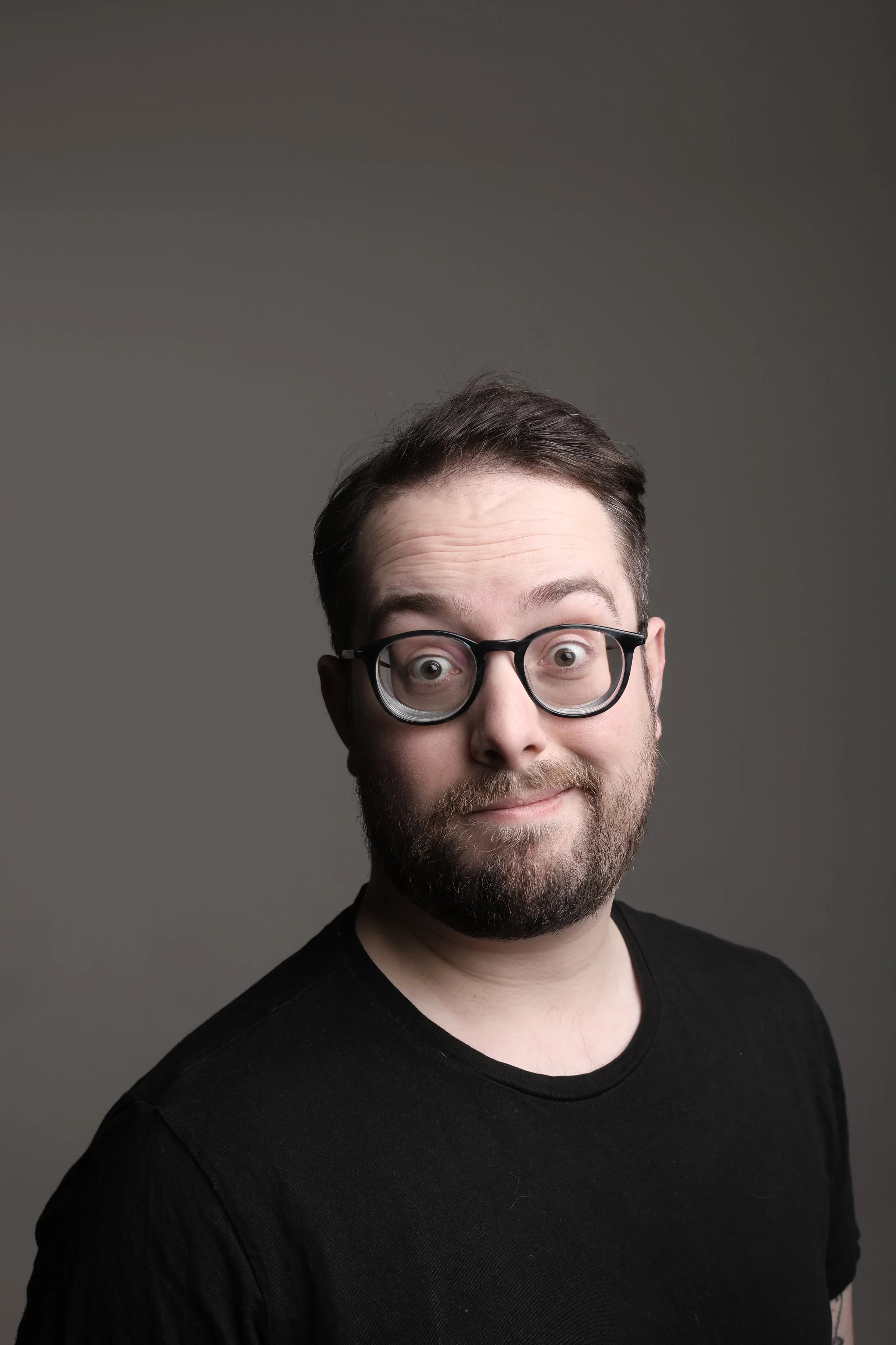 A man with glasses and a beard smiling in front of a plain gray background.