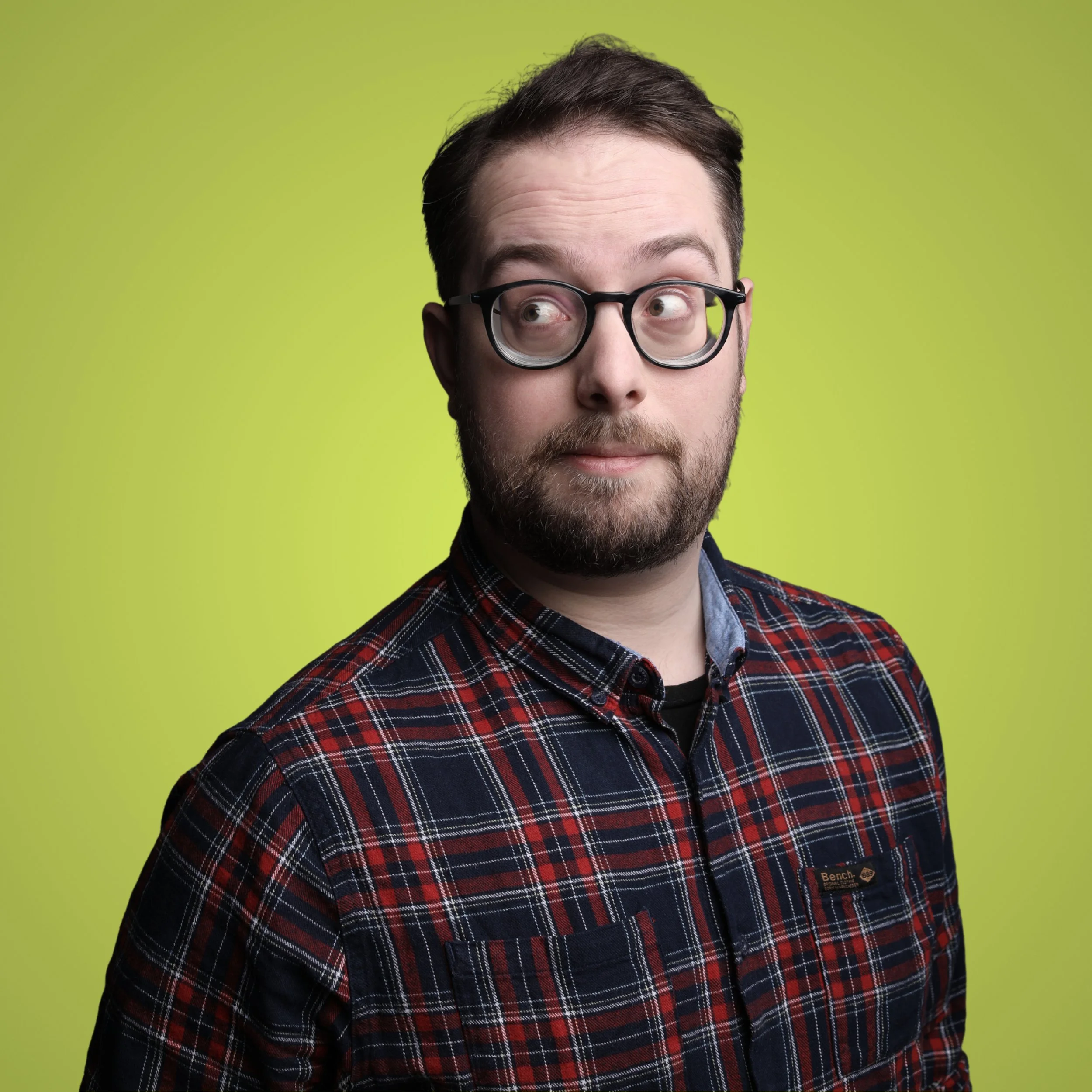 A man with glasses, a beard, and short dark hair wearing a plaid shirt against a lime green background.
