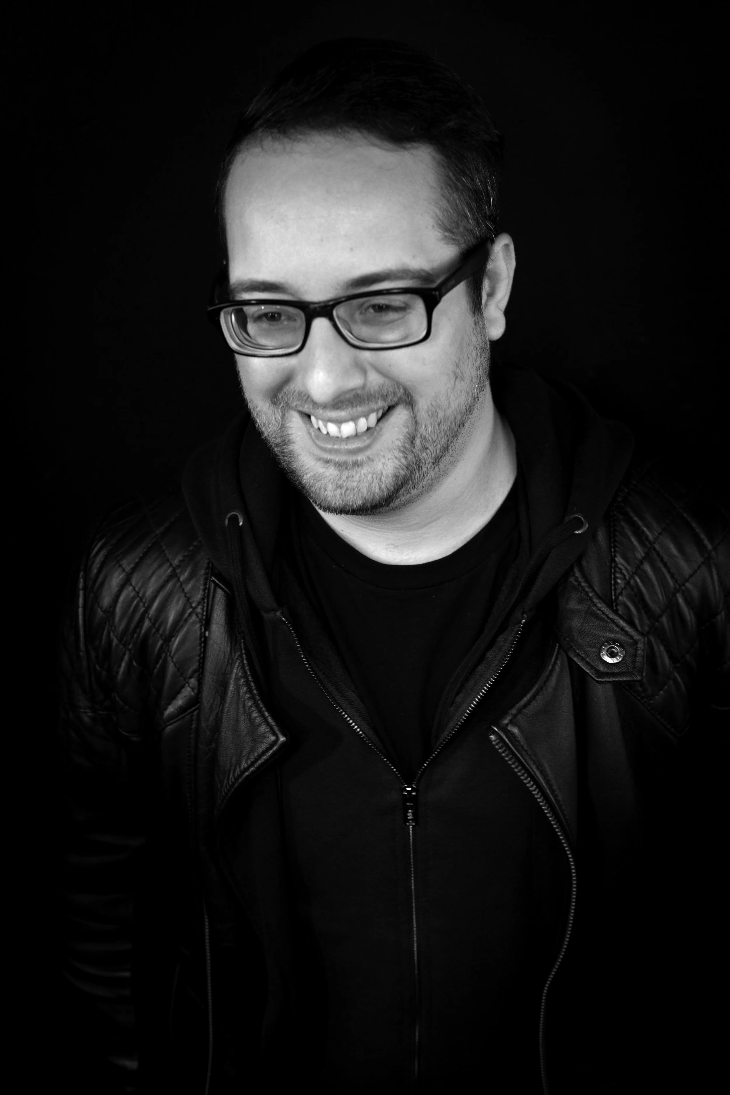 Black and white portrait of a smiling man with glasses, short dark hair, beard, wearing a leather jacket.