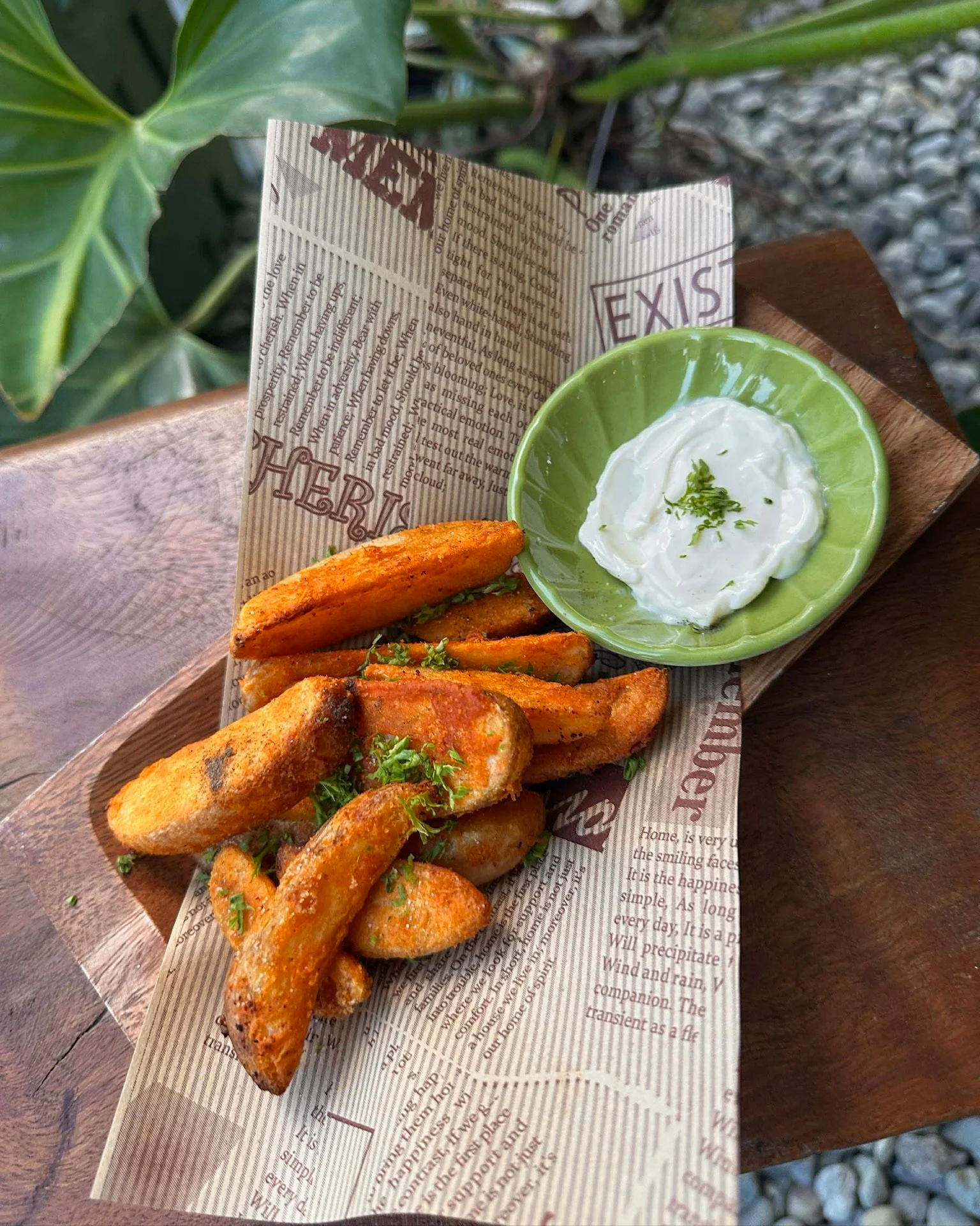 Fried potato wedges garnished with chopped parsley served with a side of creamy white dip garnished with lime zest, on a wooden tray lined with printed parchment paper, placed on a wooden table near a green plant and gravel surface.