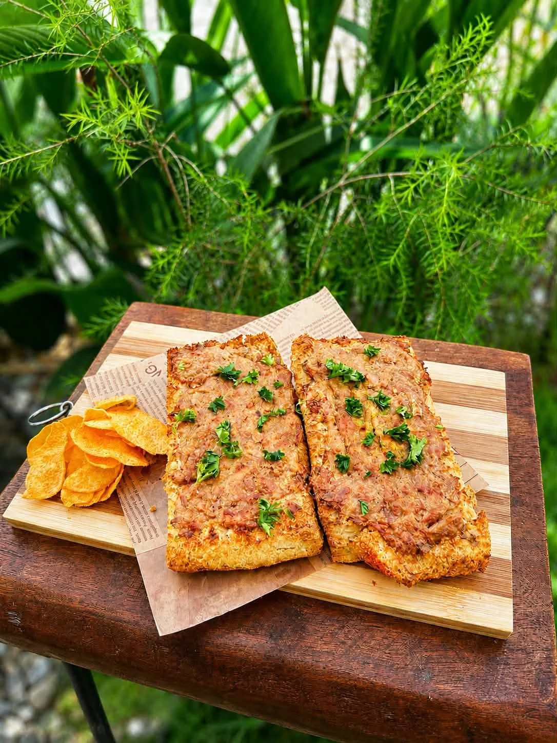 Baked fish fillets garnished with parsley on a wooden cutting board with potato chips next to it, outdoors with green plants in the background.