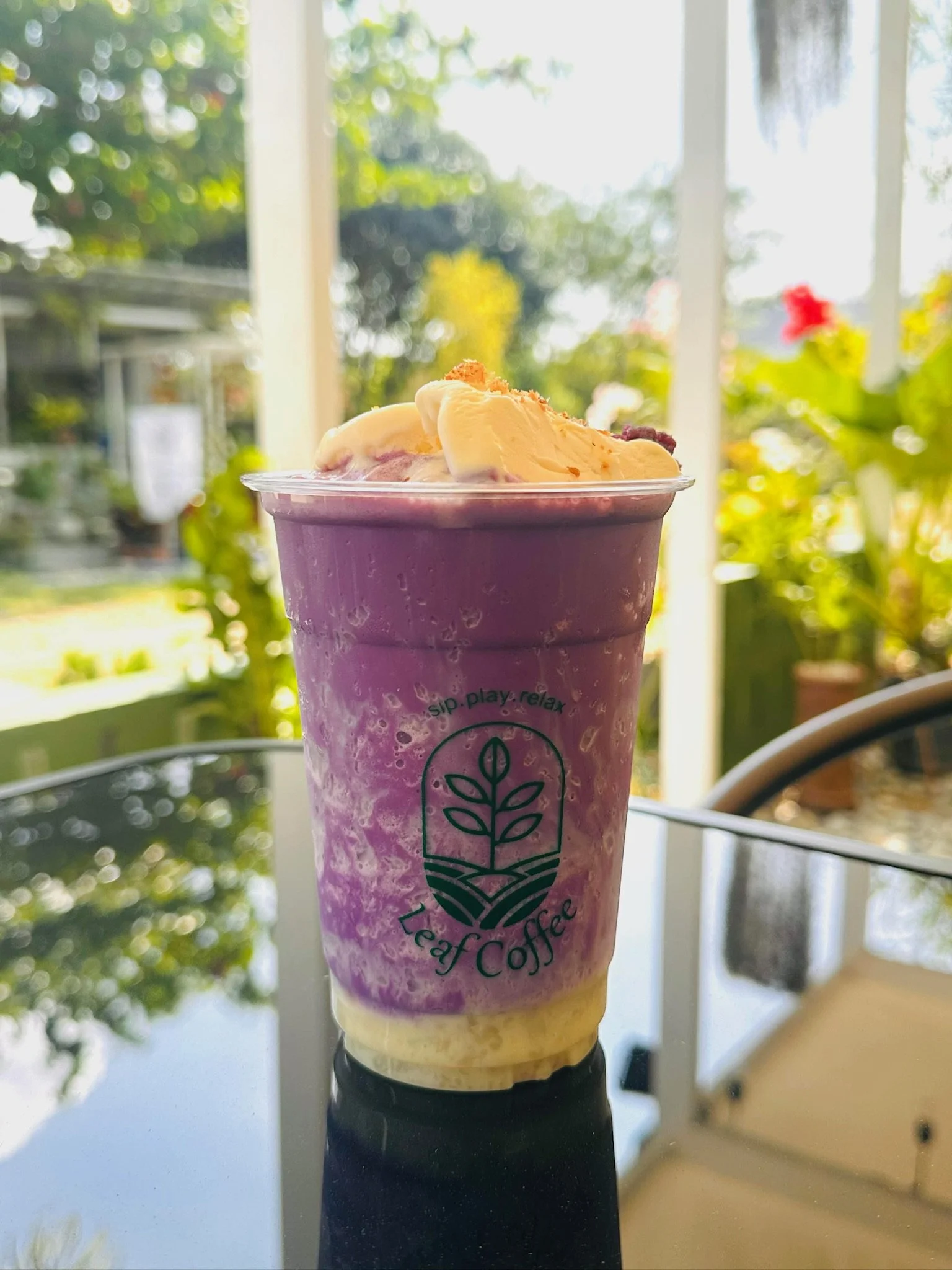 A purple blended drink in a clear plastic cup topped with vanilla soft serve ice cream and orange and red sprinkles, with a logo and text on the cup that says 'Sip, Play, Relax' and 'Tea & Coffee,' on a reflective surface with indoor and outdoor gree