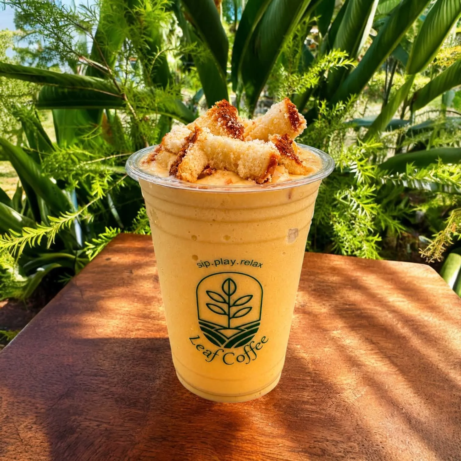 A drink in a clear plastic cup with a logo of a leaf and the words 'Leaf Coffee' and the slogan 'sip. play. relax.' is placed on a wooden table outdoors. The drink is topped with crumbled cookies and is set against a background of green plants and bl