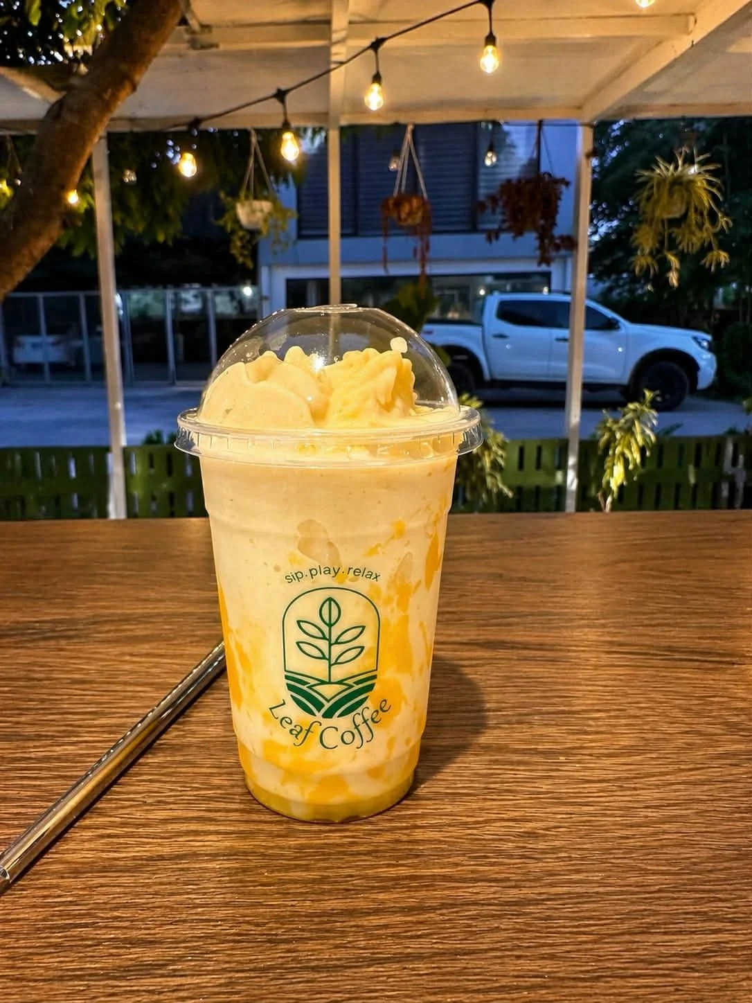 A plastic cup of mango and vanilla soft serve ice cream from Leaf Coffee on a wooden table outdoors during evening, with hanging string lights and plants in the background.