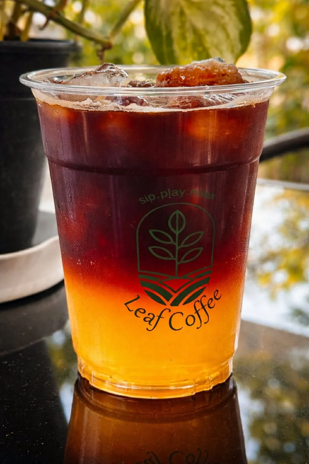 A clear plastic cup filled with iced coffee displaying the Leaf Coffee logo and the words 'sip.play.relax.' It contains ice and a layered drink with a darker upper layer and a lighter yellow-orange bottom layer.