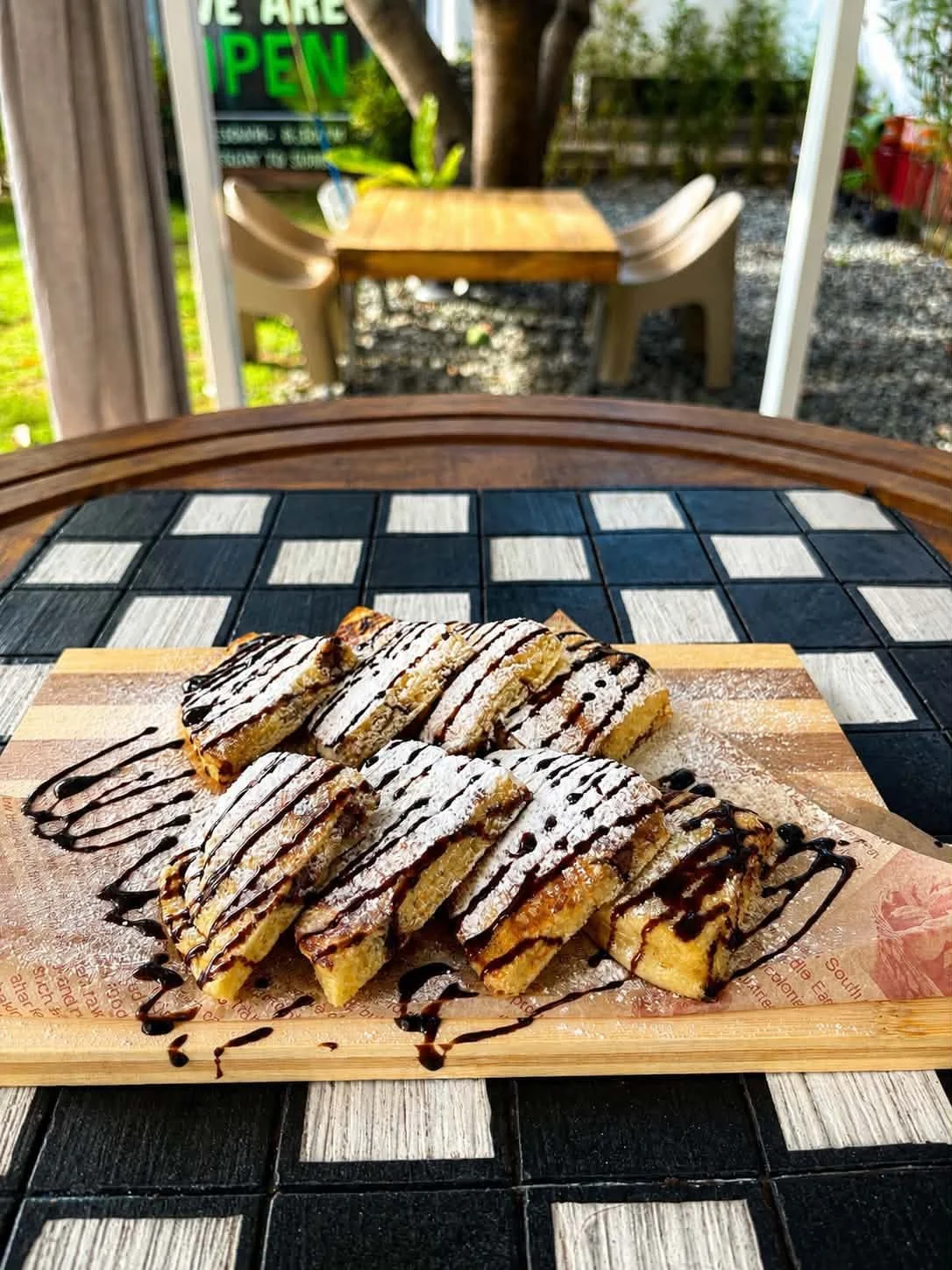 Chocolate-drizzled dessert bars on a wooden serving board, dusted with powdered sugar, on a checkered black and white table outdoors.