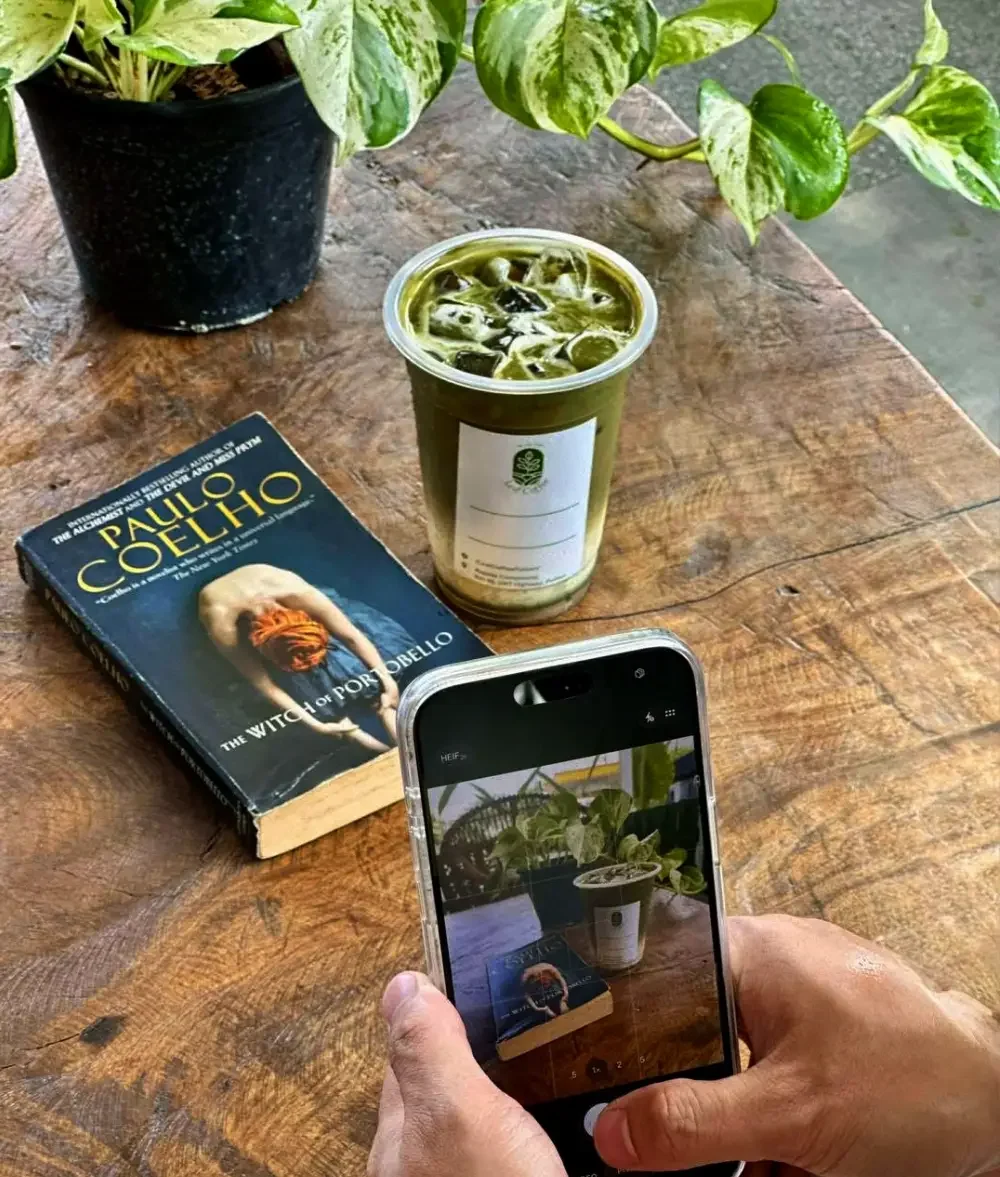 A wooden table with a book titled 'Paulo Coelho,' a cup of iced coffee, a potted plant, and a person taking a photo of the scene with a smartphone.