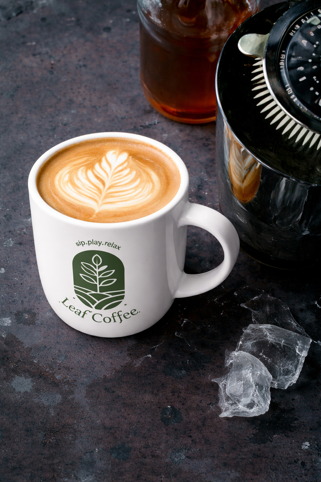 A white coffee mug with the Leaf Coffee logo contains a latte with latte art on top, placed on a dark surface with ice cubes nearby, along with a glass of iced tea and a coffee grinder.