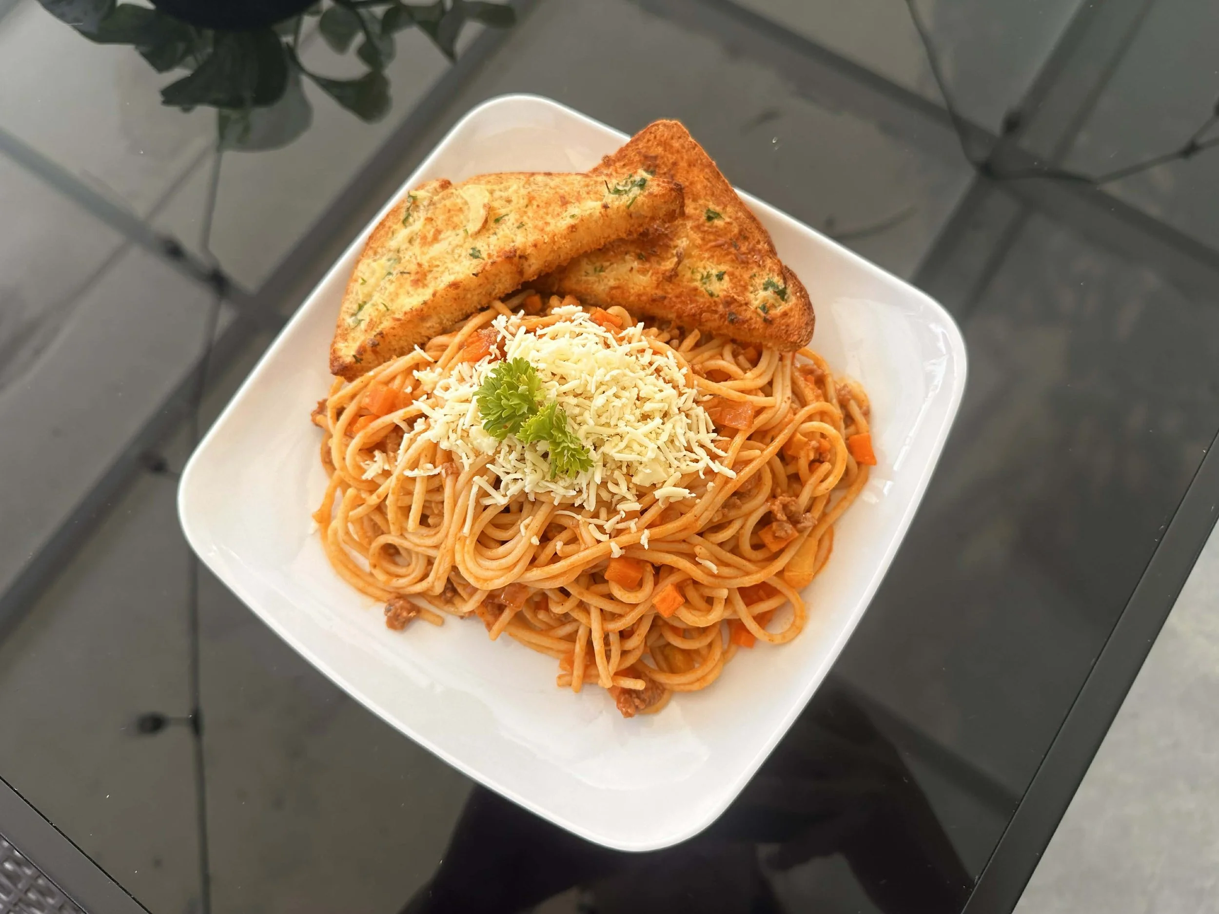 Plate of spaghetti with tomato sauce, grated cheese, parsley, and two slices of garlic bread on a white square plate placed on a black table.