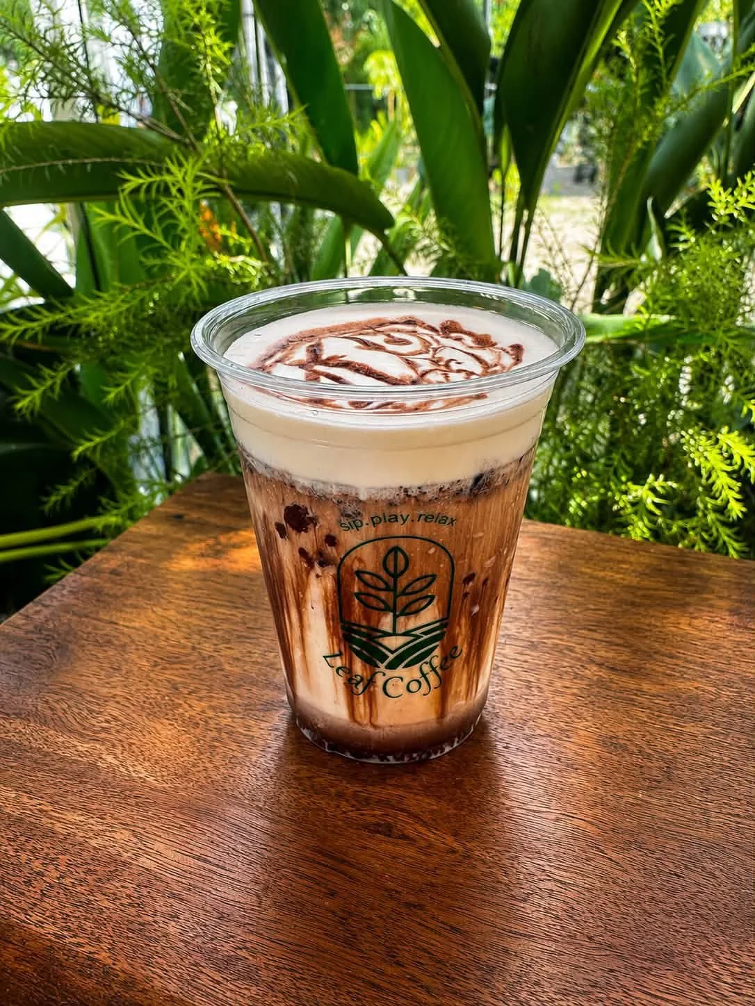 A plastic cup filled with a layered iced coffee topped with cream and a drizzle of chocolate syrup, placed on a wooden table with green foliage in the background.
