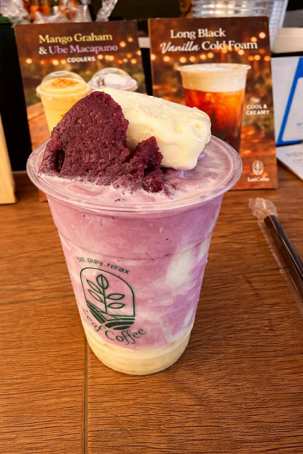 A pink and purple swirled frozen drink in a clear plastic cup topped with vanilla ice cream and a chunk of red velvet cake. In the background are two menu boards advertising mango graham and vanilla cold foam drinks.