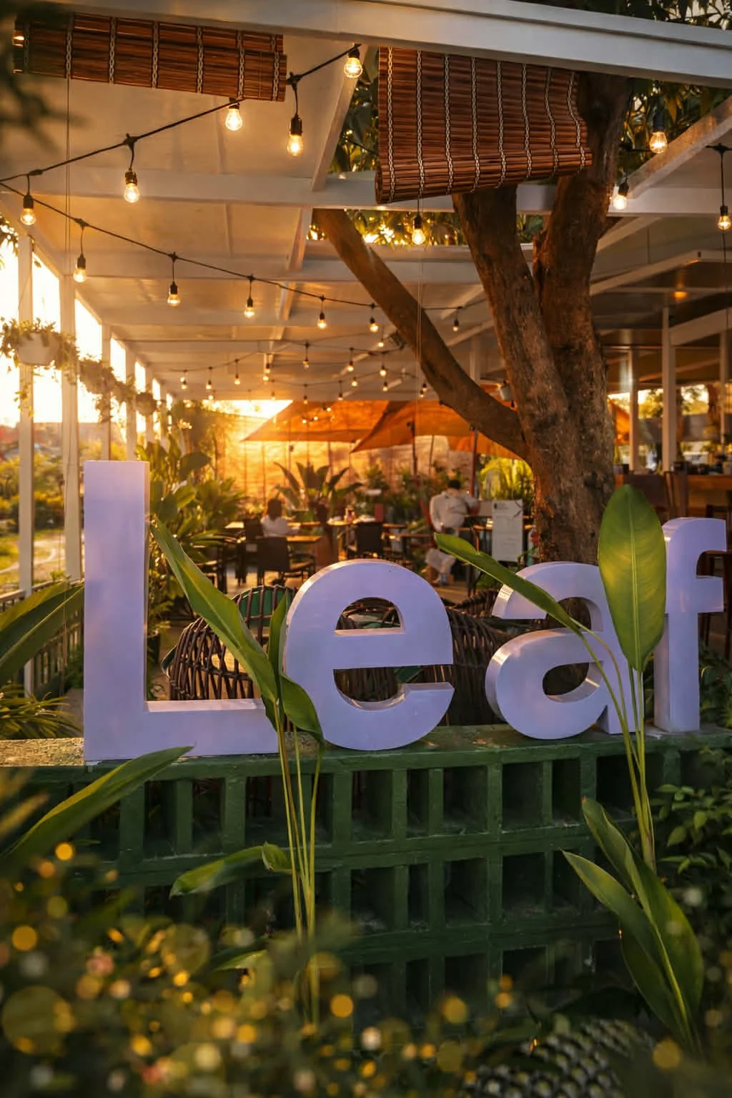 Sunset view of a cozy outdoor restaurant or cafe with string lights, a large tree, and a decorative 'Leaf' sign in the foreground, lush green plants, and dining tables with umbrellas in the background.