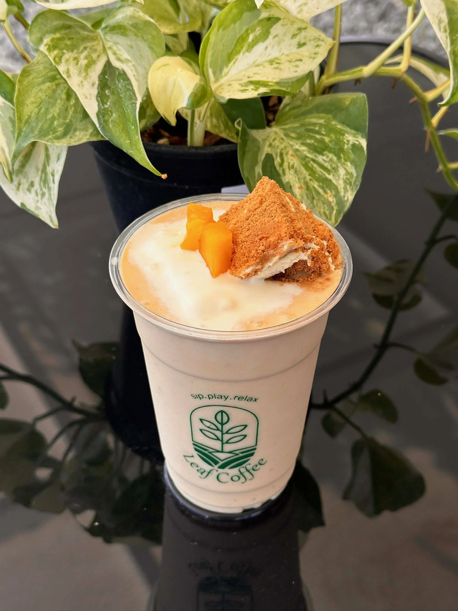 A cup of iced coffee with whipped cream, topped with a small piece of mango and a sprinkle of cinnamon, placed on a black surface with a potted variegated green and white leaf plant in the background.