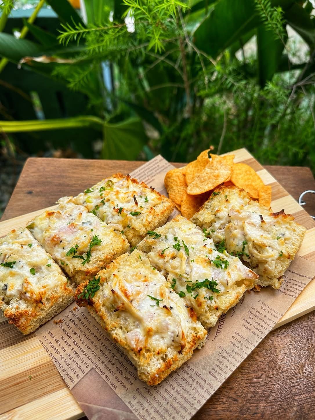 Baked cheesy bread slices garnished with herbs on a wooden serving board with potato chips in the background.