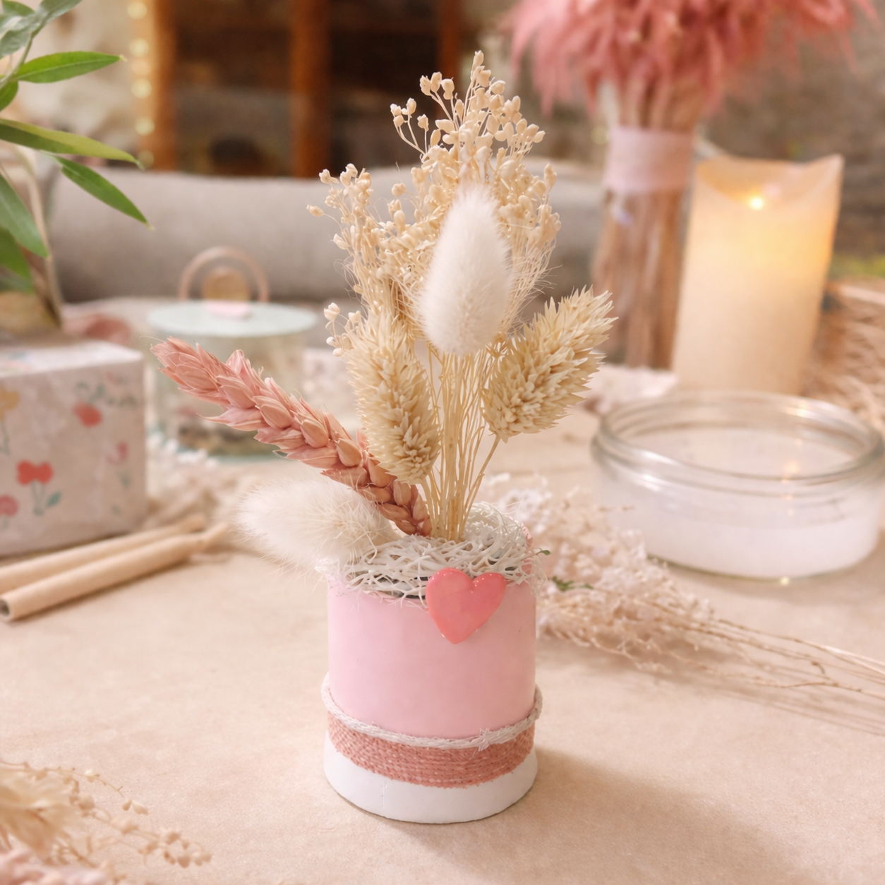 Une décoration florale en rose pastel avec des fleurs séchées dans un petit vase, entourée d'éléments décoratifs et de bougies