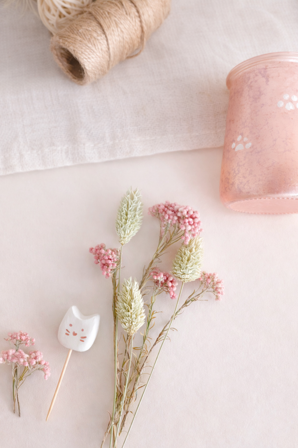 Fleurs roses et blanches, un marqueur en forme de chat, une pelote de laine beige, un pot en verre rose, et un tissu blanc