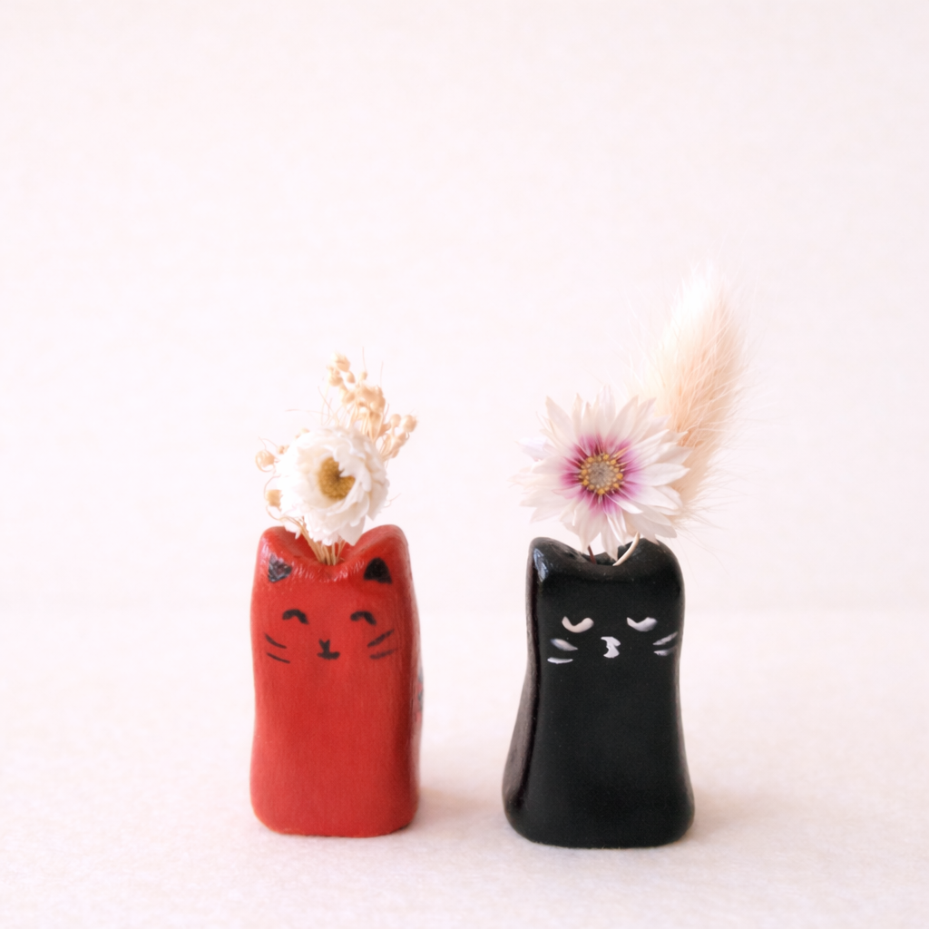 Deux vases en forme de chats avec des fleurs dans dessus, un rouge et un noir, sur fond blanc.