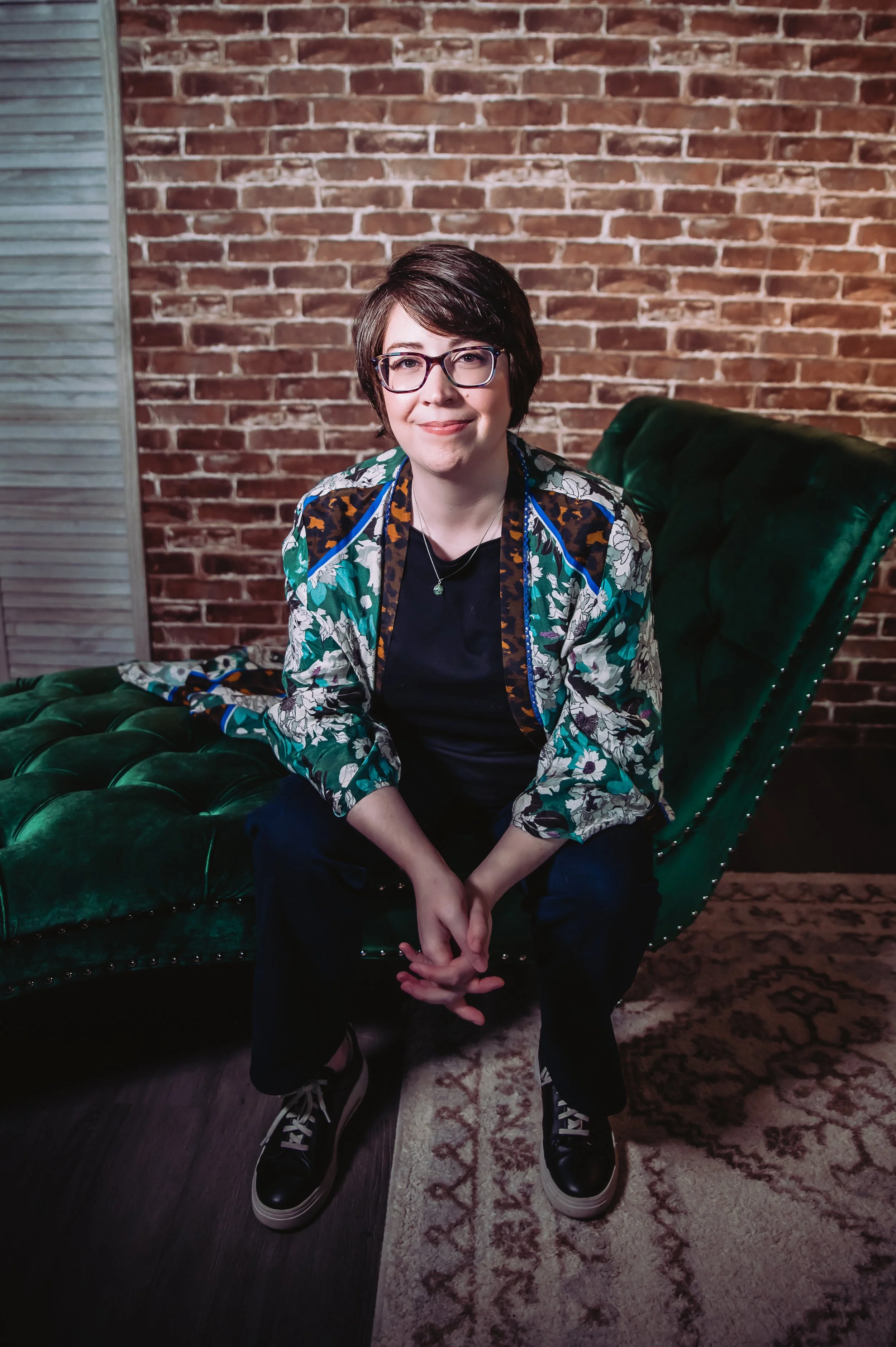 A young woman with short dark hair, glasses, and a patterned jacket sits on a green velvet couch with a brick wall background. She has a subtle smile and is wearing black pants and sneakers, with her hands clasped in her lap.