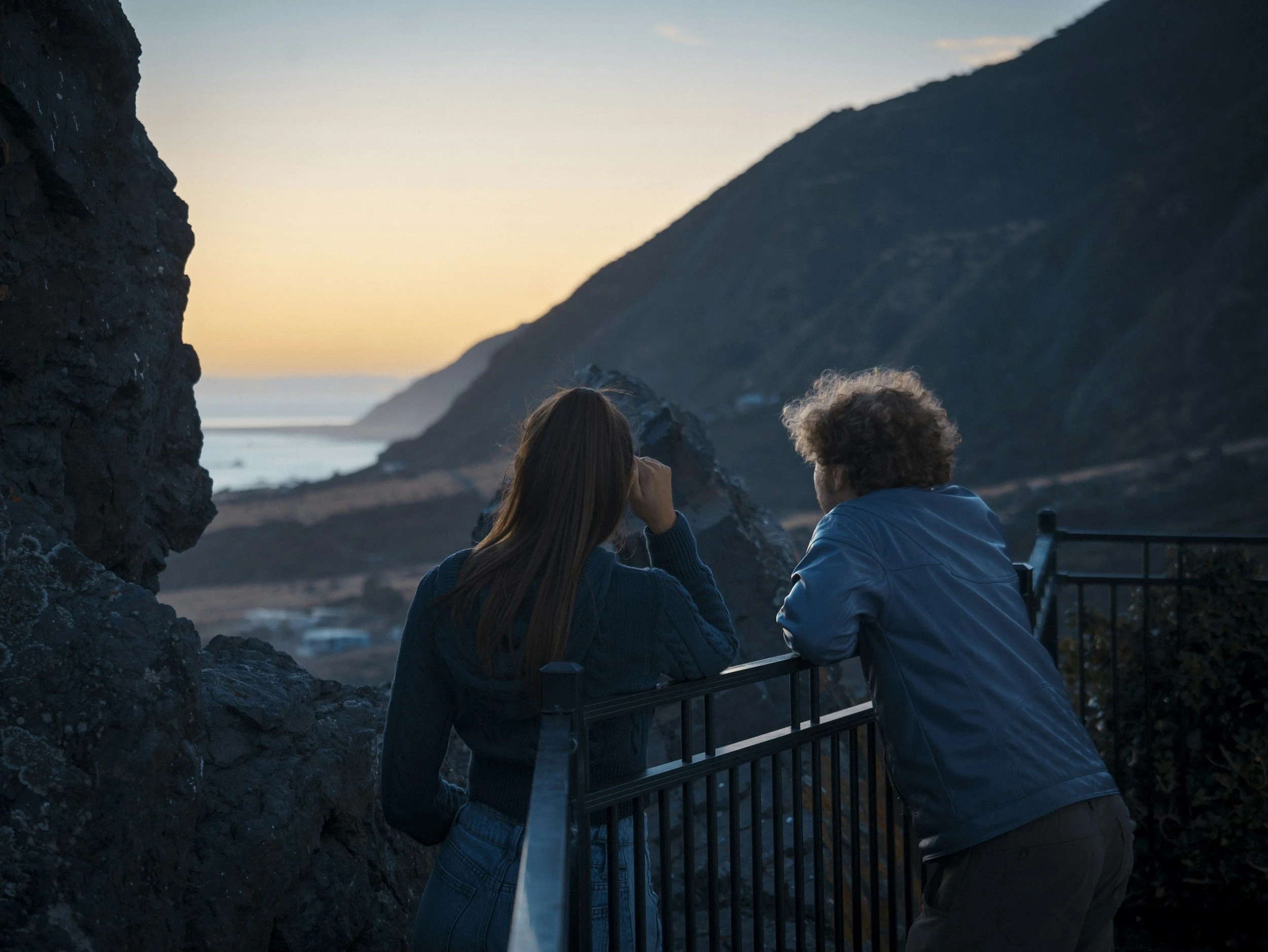 Two people with dark hair, one with curly hair, are sitting on a bench by the coast during sunset, looking at the ocean and hills in the distance.
