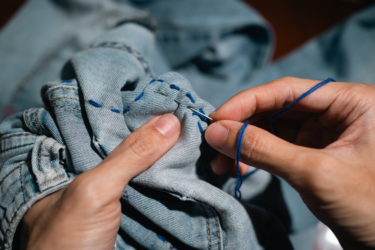 Close-up of hands sewing denim jeans with a needle and blue thread.