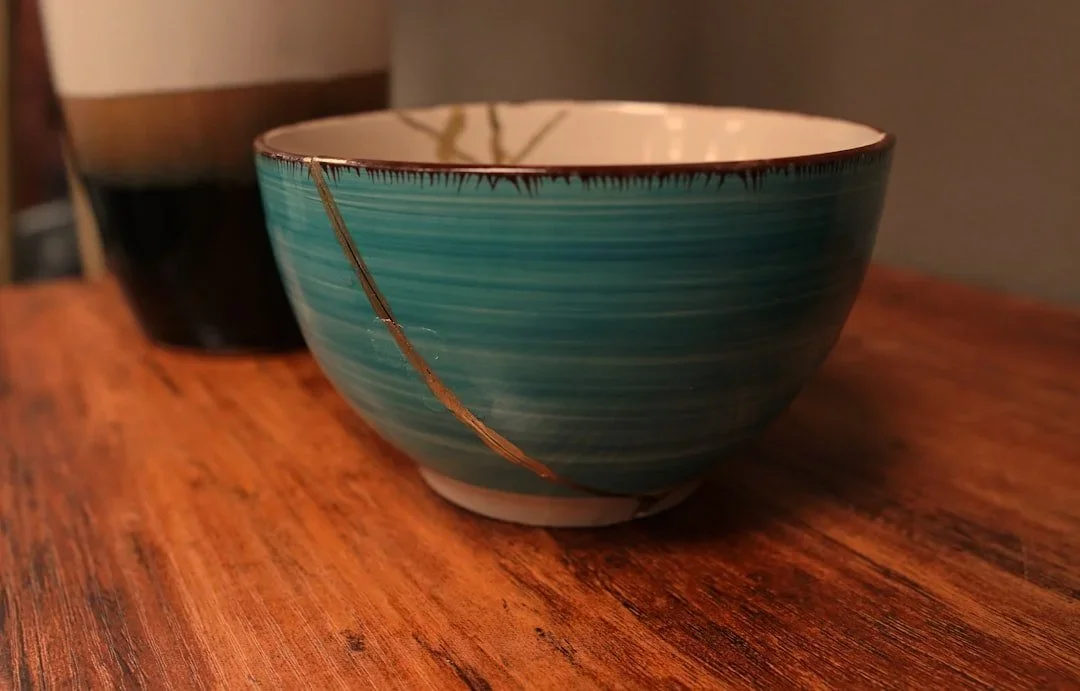 A ceramic bowl with a teal and white swirl pattern and kintsugi, placed on a wooden surface with a dark vase in the background.