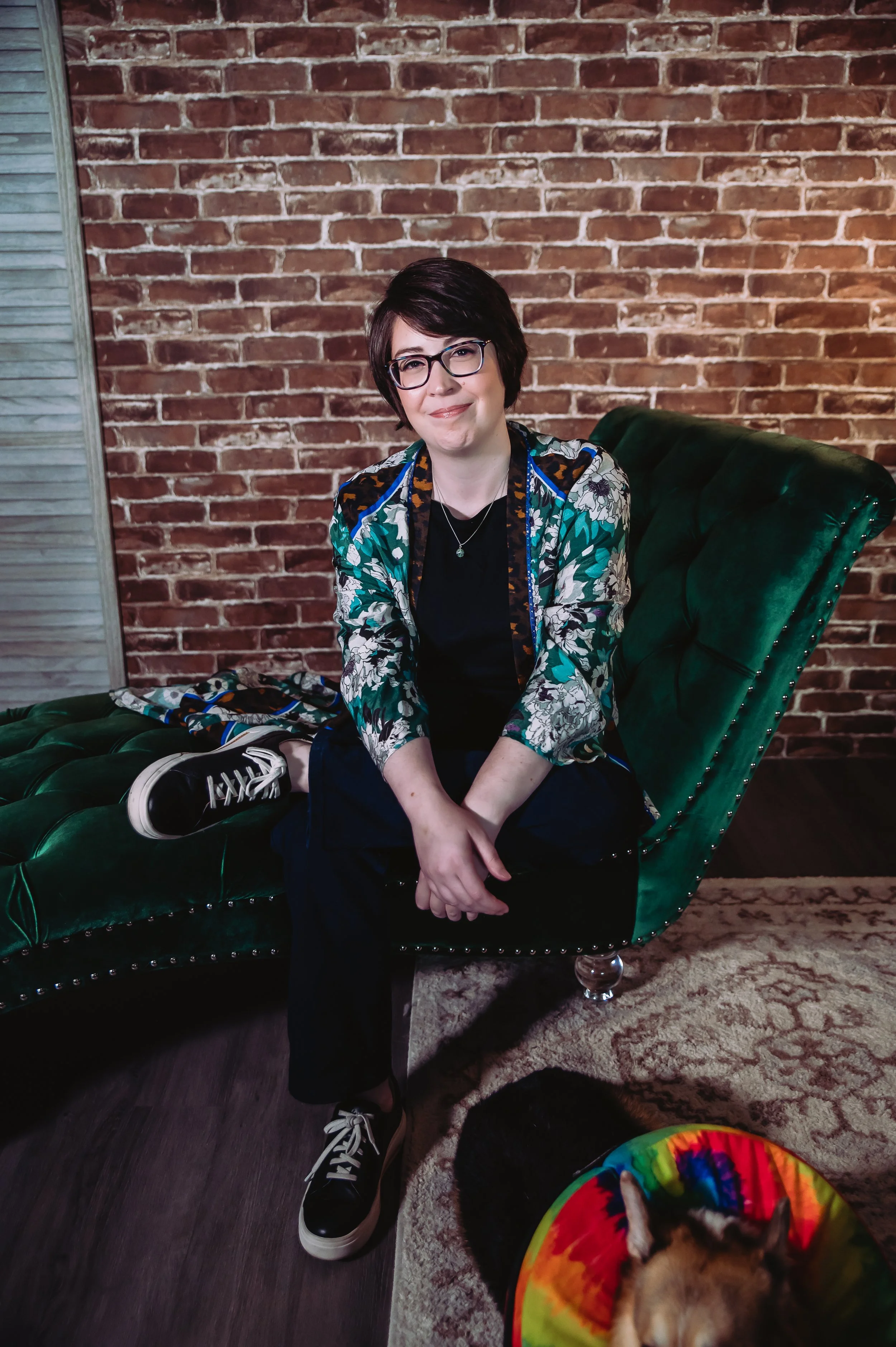 A woman with short dark hair, glasses, and a floral jacket sitting on a green velvety couch in front of a brick wall, with her legs crossed and smiling at the camera. A dog wearing a colorful tie-dye cone is lying on the floor near her, partially visible.