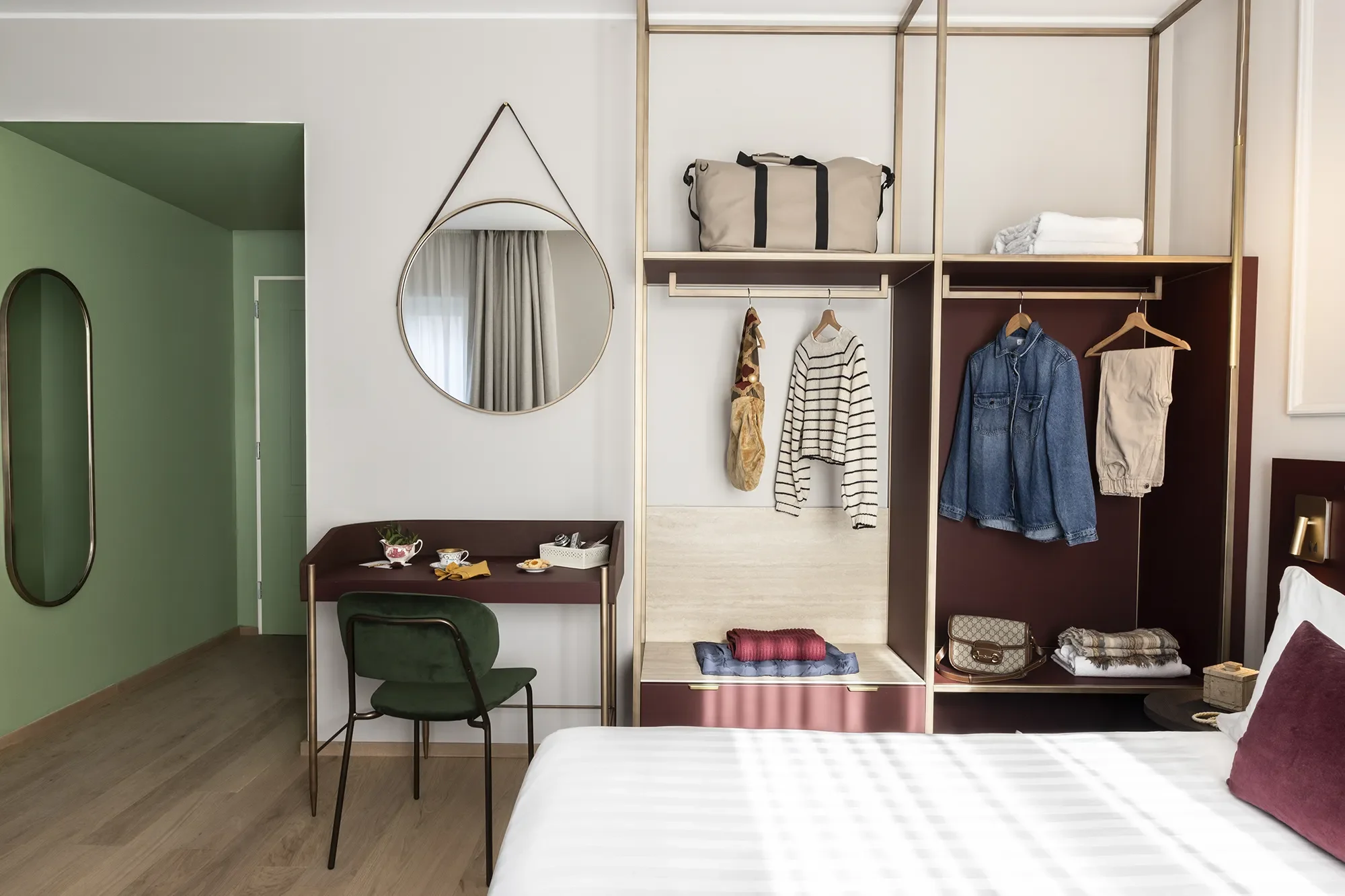 Interno di una camera con letto, armadio aperto con vestiti e oggetti, specchio tondo, scrivania con sedia, pareti di colore bianco e verde, pavimento in legno.