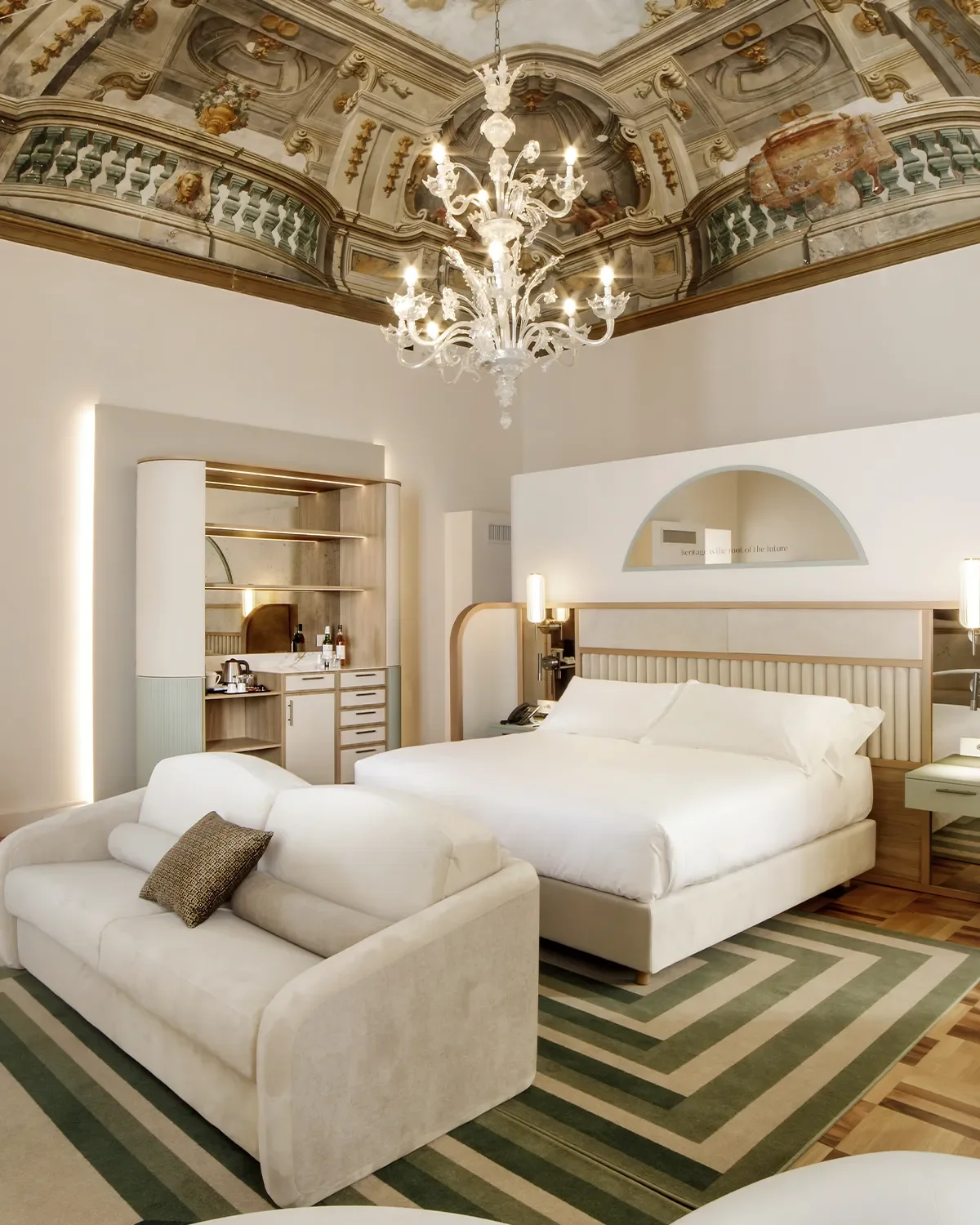 Camera da letto elegante con un grande letto con lenzuola bianche, un divano beige con un cuscino, un tappeto con motivi geometrici verdi e beige, e un'illuminazione morbida. Sopra, un soffitto decorato e un lampadario in cristallo.