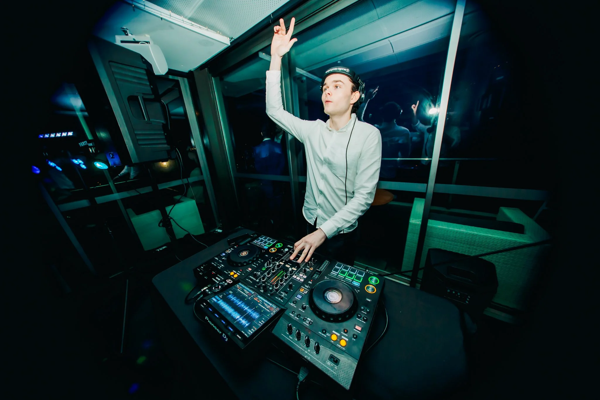 A DJ wearing headphones is raising one hand in the air while operating a DJ controller and mixer at a nightclub.