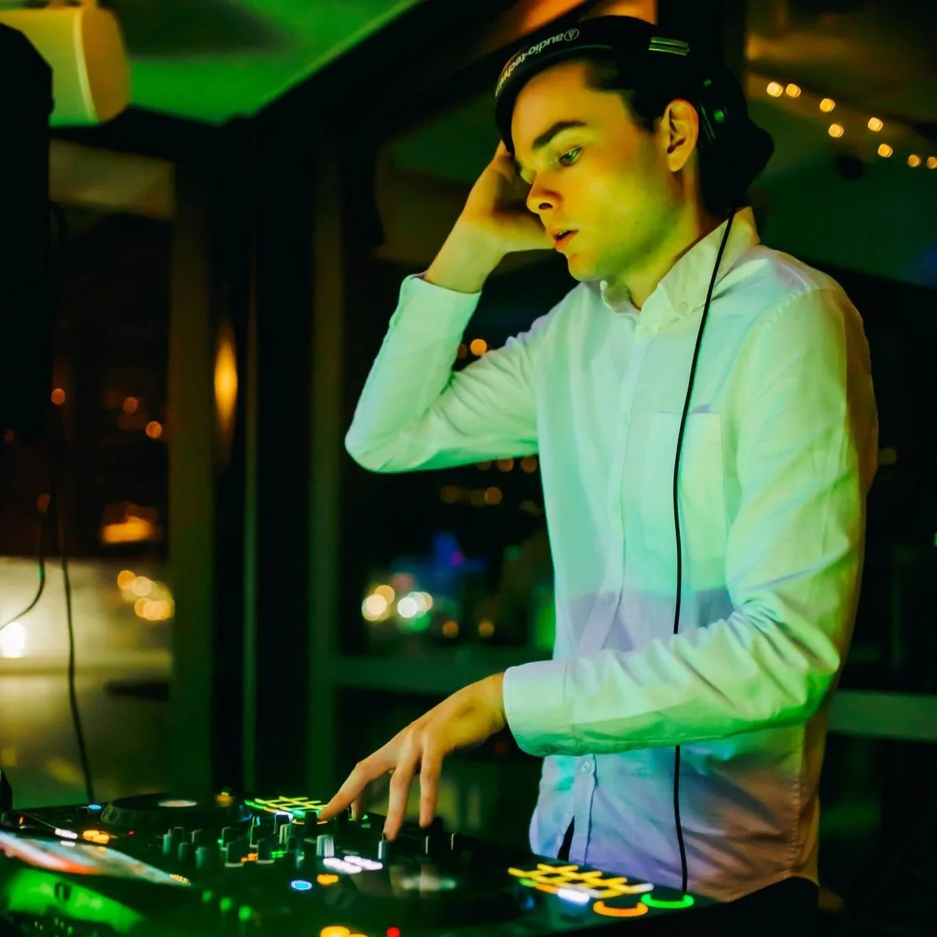 DJ mixing music at a nightclub with a focus on the DJ's hands and equipment, wearing headphones and a white shirt, illuminated by colorful lights.