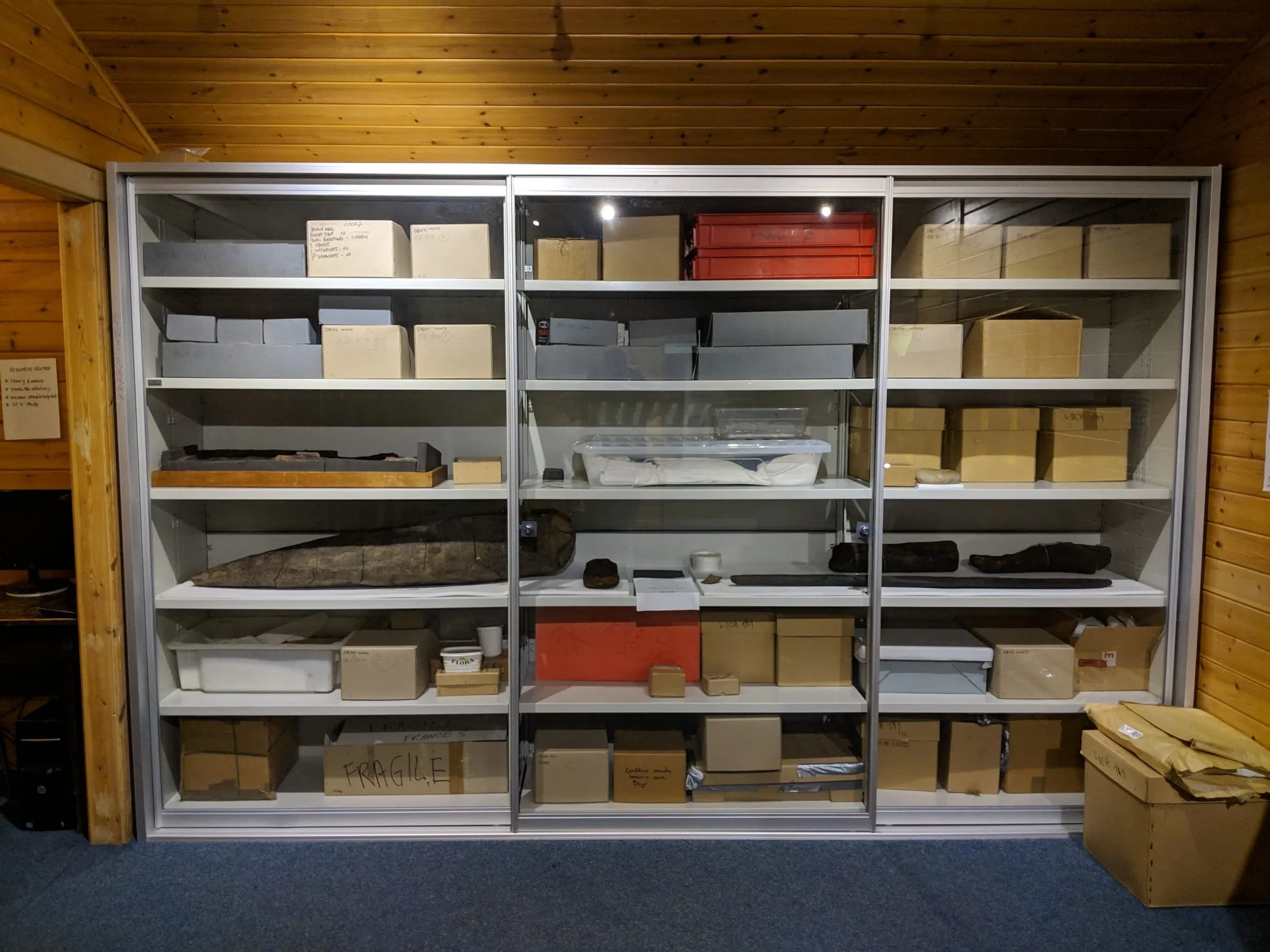 A metal shelving unit with glass doors containing various cardboard boxes, some labeled, and artifacts, in a wooden room.
