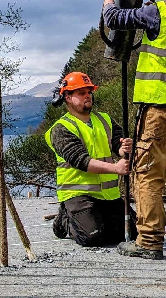 Worker kneeling on the ground wearing a yellow safety vest and a hard hat, working with a large drill or tool on a construction site outdoors, with trees and mountains in the background.