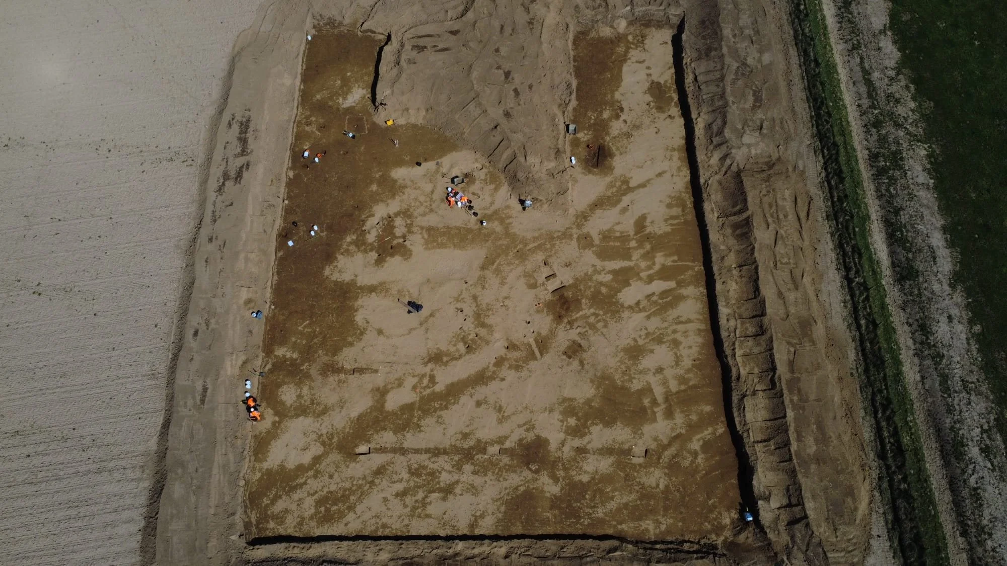 An aerial view of an archaeological excavation site with several workers and tools scattered across the area, surrounded by tilled field and green land.