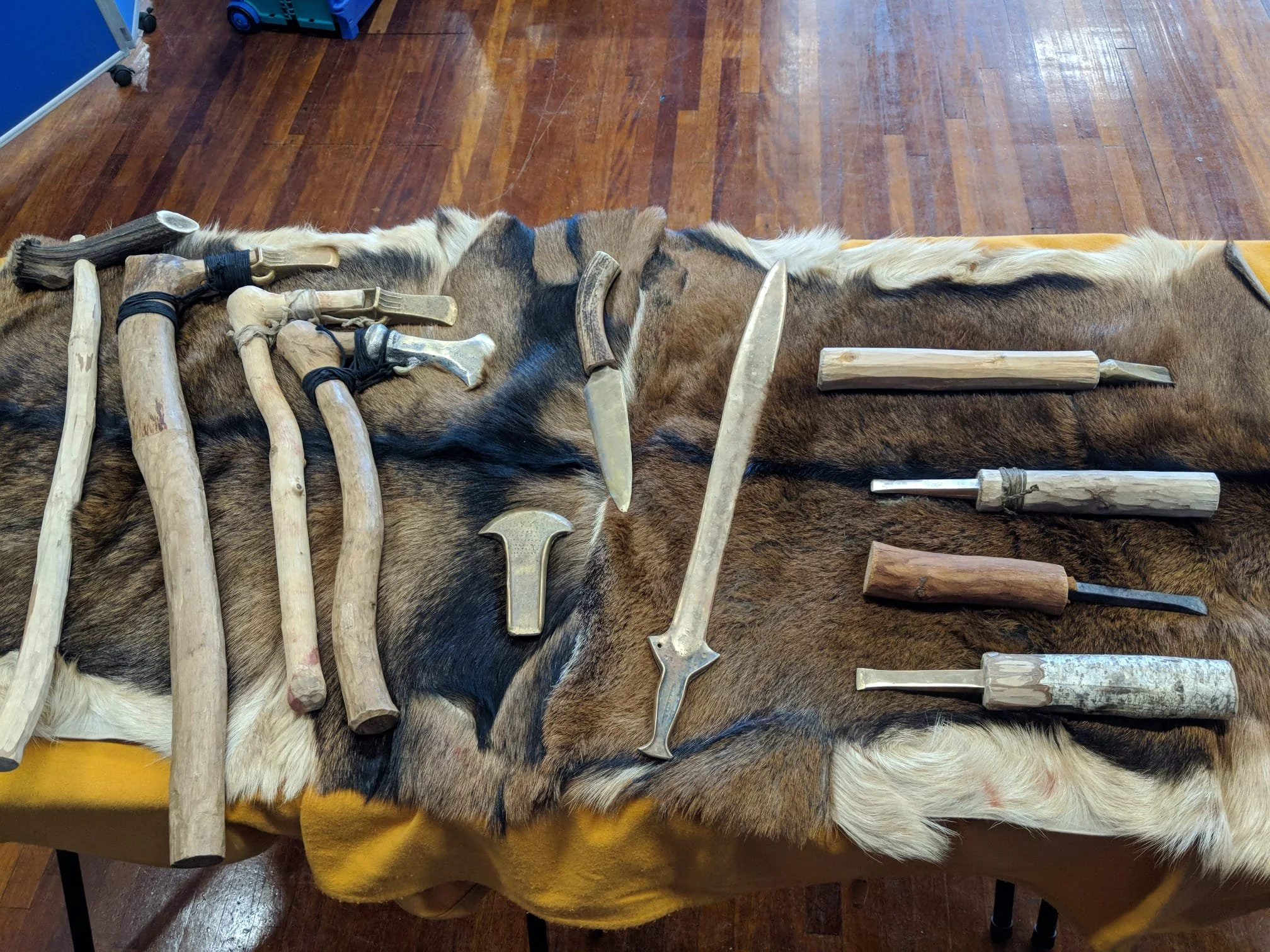 Collection of antique and rustic knives and tools made of wood and metal, laid out on a cowhide rug on a wooden floor.