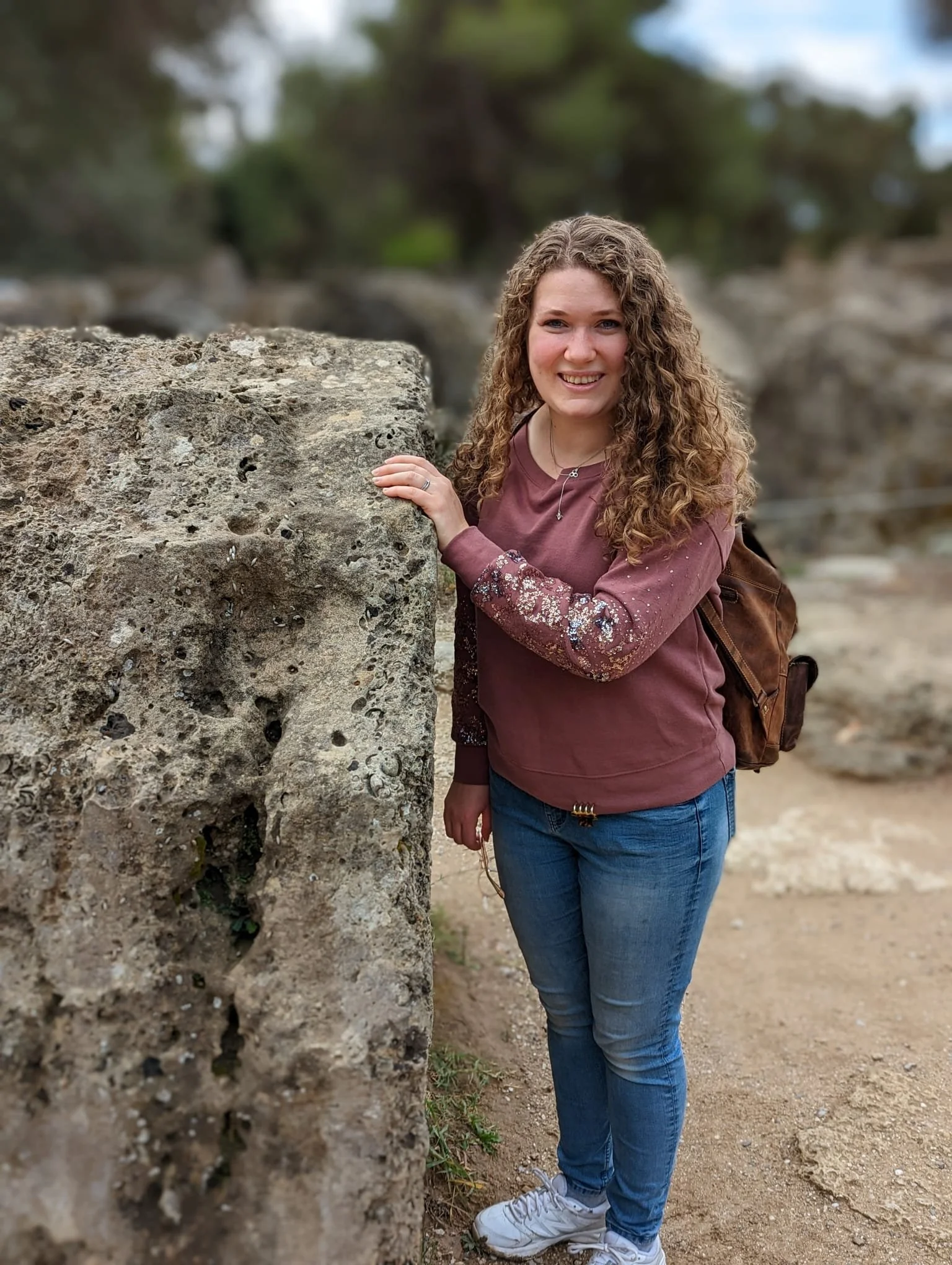 A young woman with curly hair smiling outdoors, wearing a maroon sweatshirt with embellished sleeves, blue jeans, white sneakers, and a brown backpack, standing next to a large textured rock.