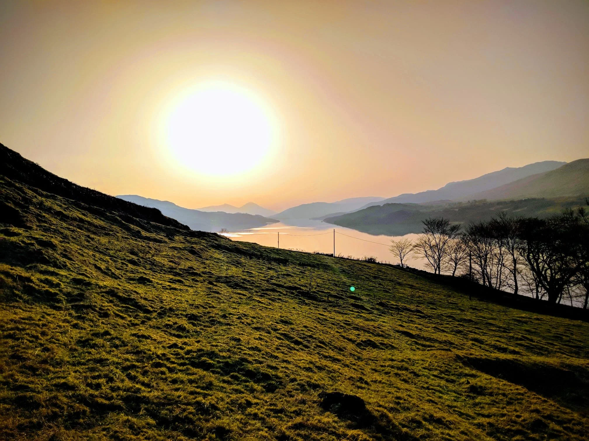 Landscape with grassy hill in the foreground, leafless trees to the right, a wide river or lake reflecting the sky, and distant mountains under a setting or rising sun in the sky.