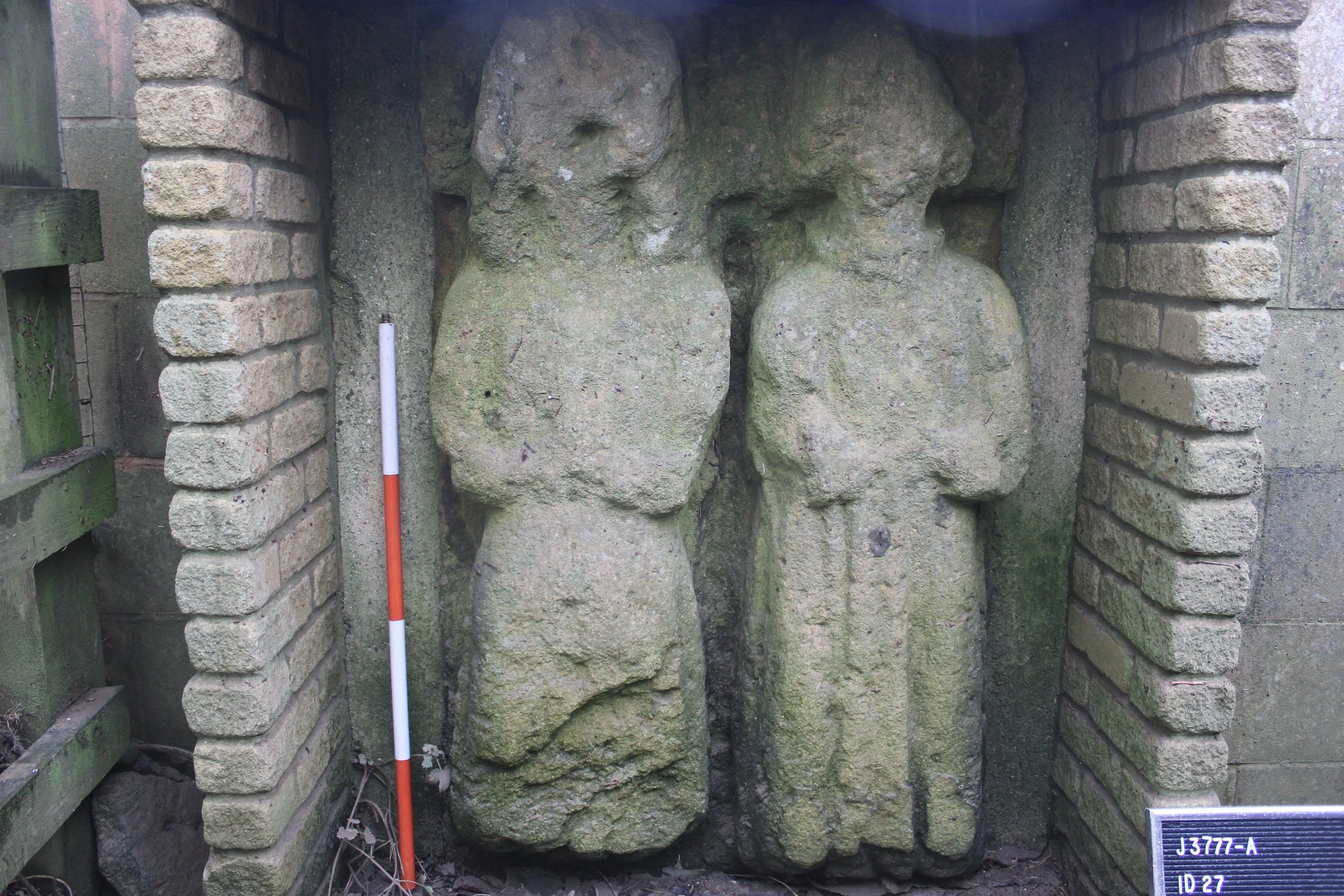 Ancient stone sculpture of two human figures carved into a large, weathered rock, flanked by brick walls, with an identification plaque in the lower right corner.