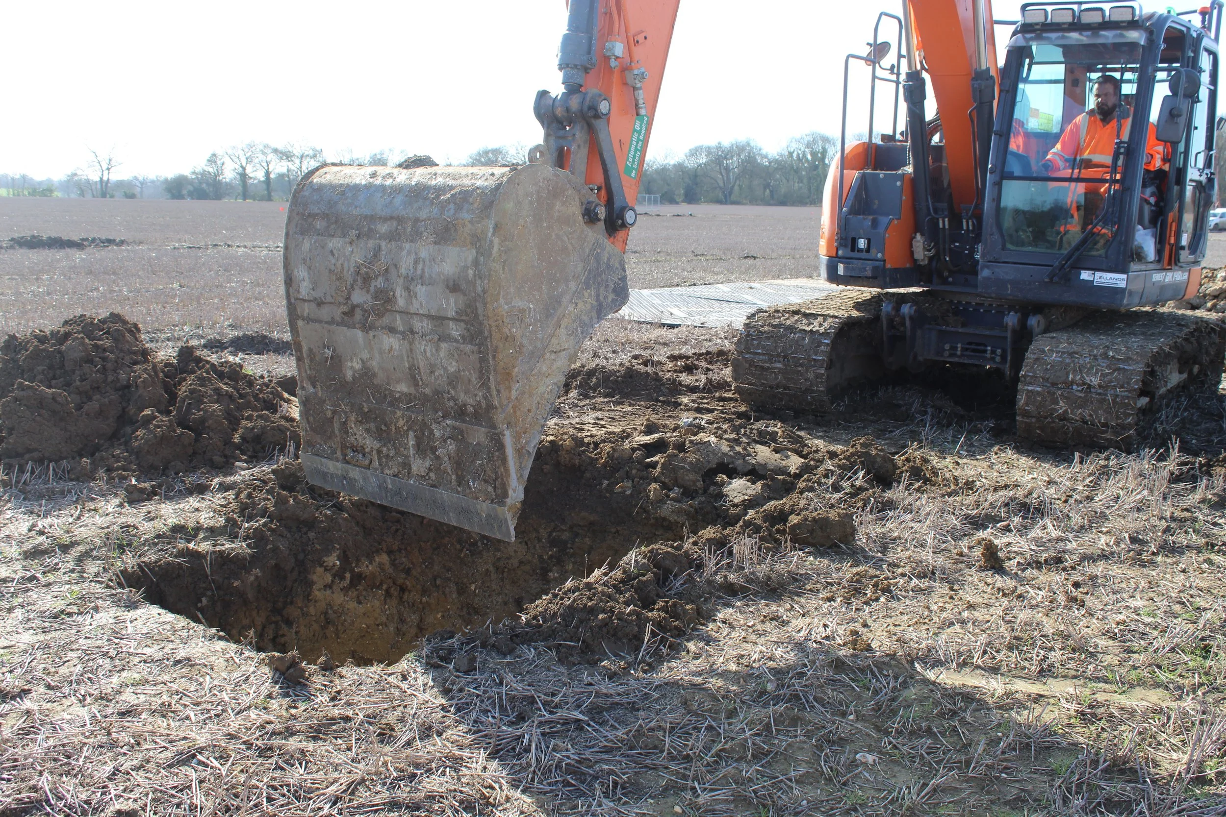 An excavator with an orange arm and black tracks digging a hole in a field with dry grass and soil, with a worker in an orange safety vest operating it.