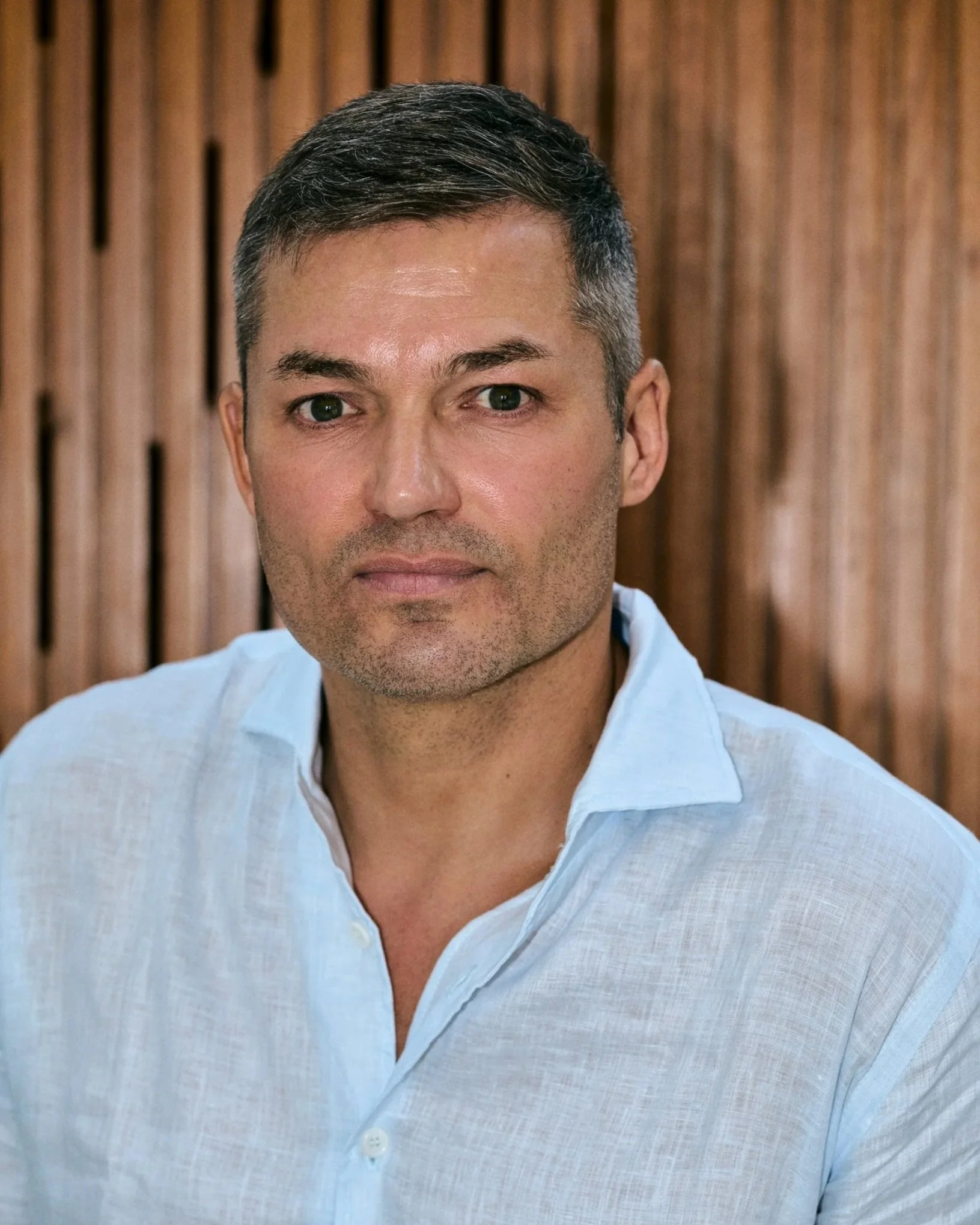 A portrait of a man with short dark hair, wearing a light blue collared shirt, in front of a wooden background.
