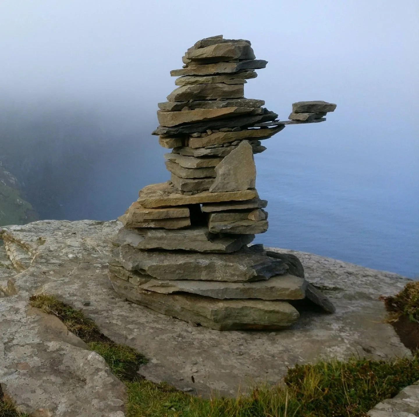 Stacked flat stones balanced on top of each other on a rocky surface near the water with misty hills in the background.