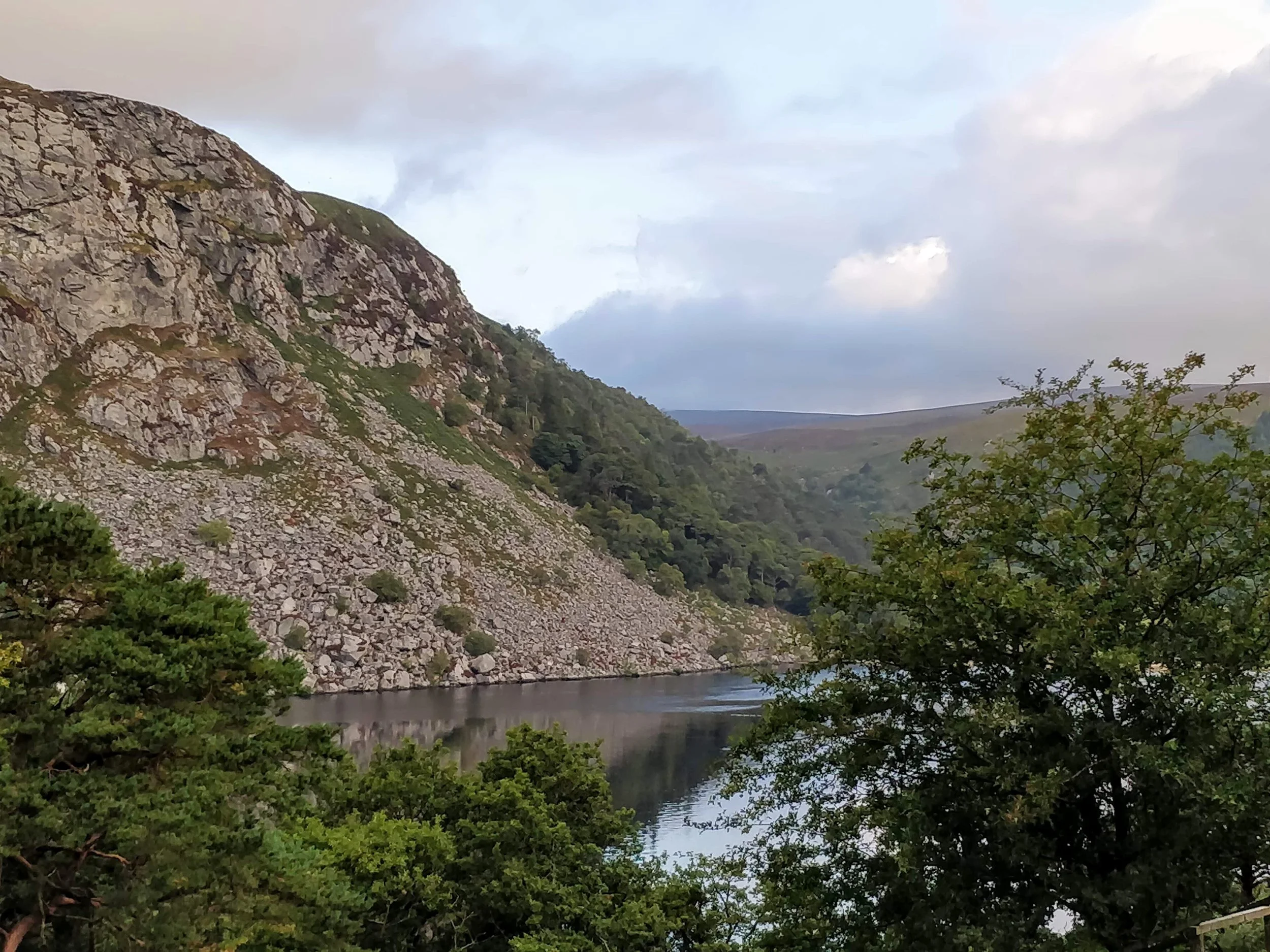 A scenic view of a river flowing through a valley surrounded by rocky and green hills under a cloudy sky.