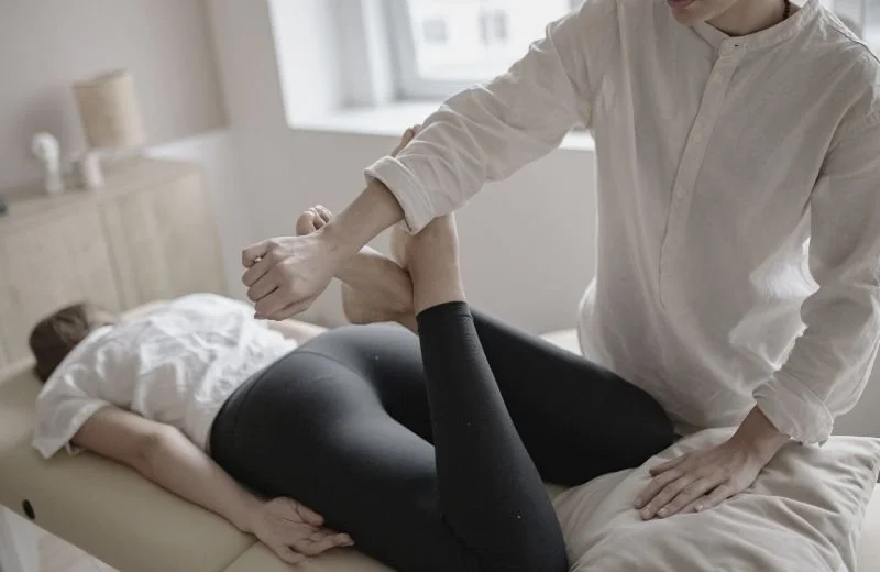 A person receiving Amatsu therapy from another person on a massage table in a bright, minimalistic room.