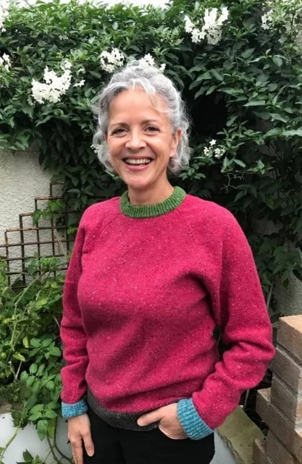 A smiling woman with curly grey hair, wearing a pink sweater with green accents, standing in front of green foliage and white flowers in a garden.