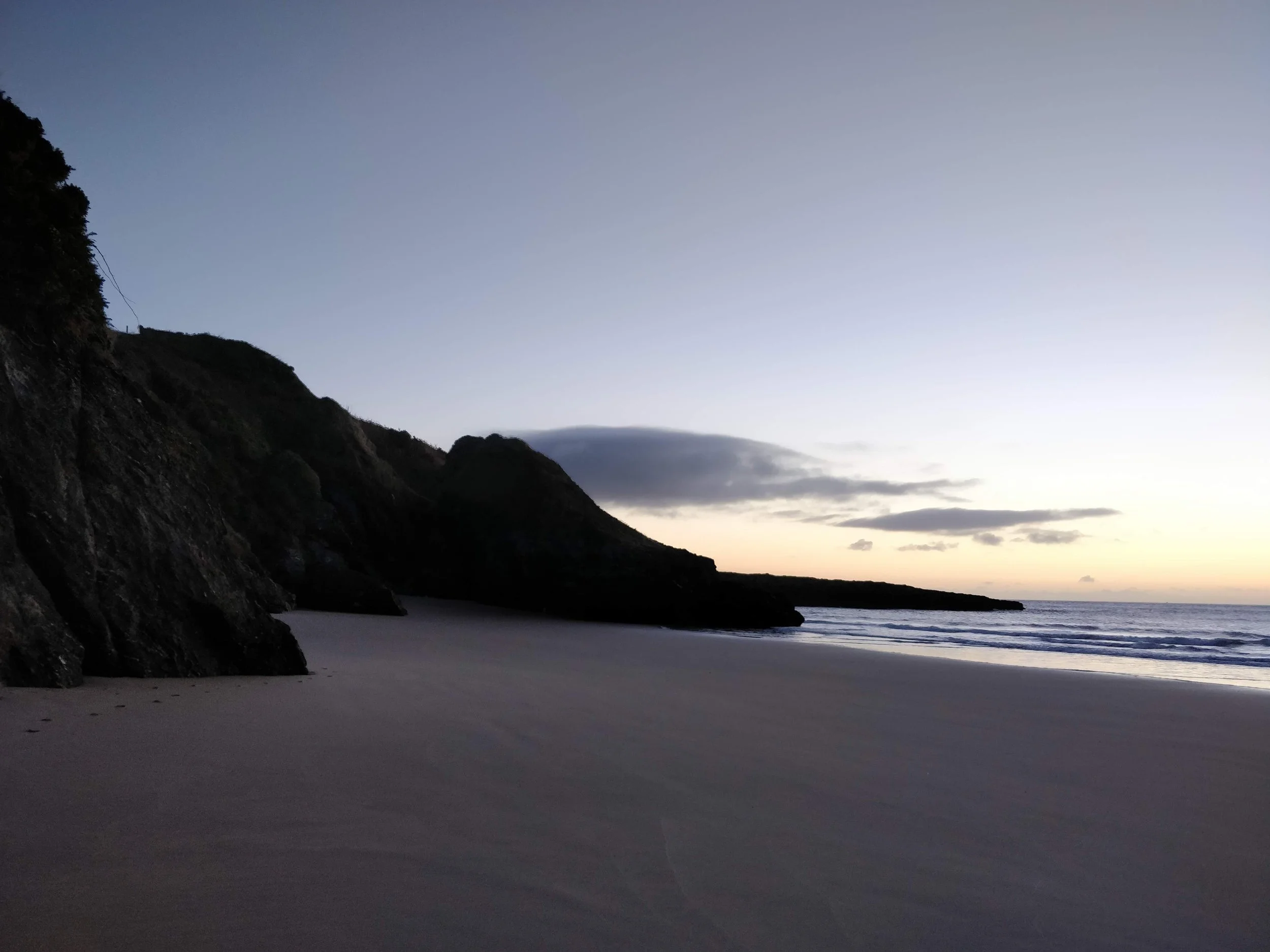 A peaceful beach scene at sunset with a sandy shore, calm waves, and large rocky cliffs on the left side under a sky with some clouds.